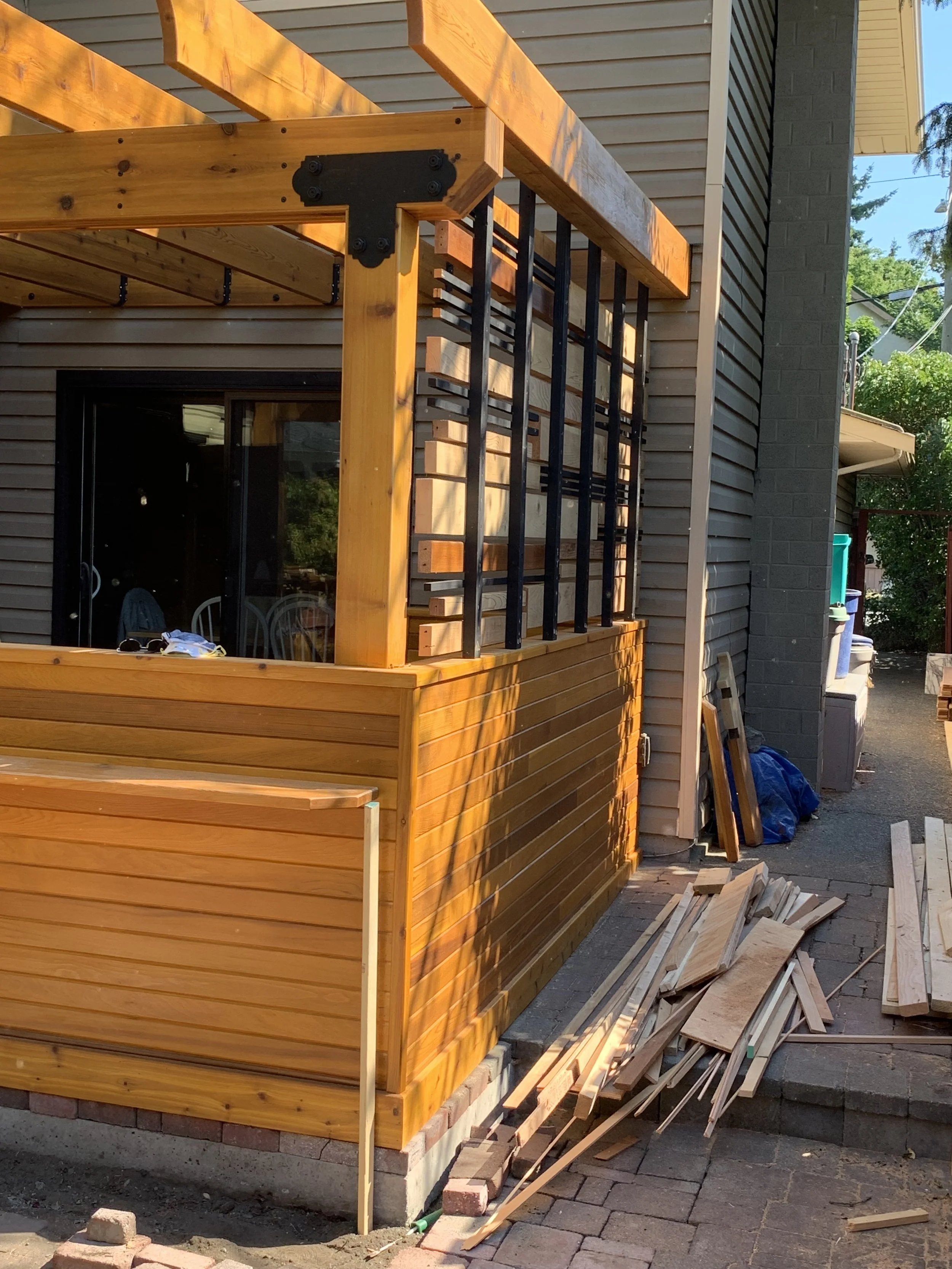 Wooden deck construction with unfinished wood and black metal brackets on the side of a house, with tools, wood planks, and construction materials scattered around.