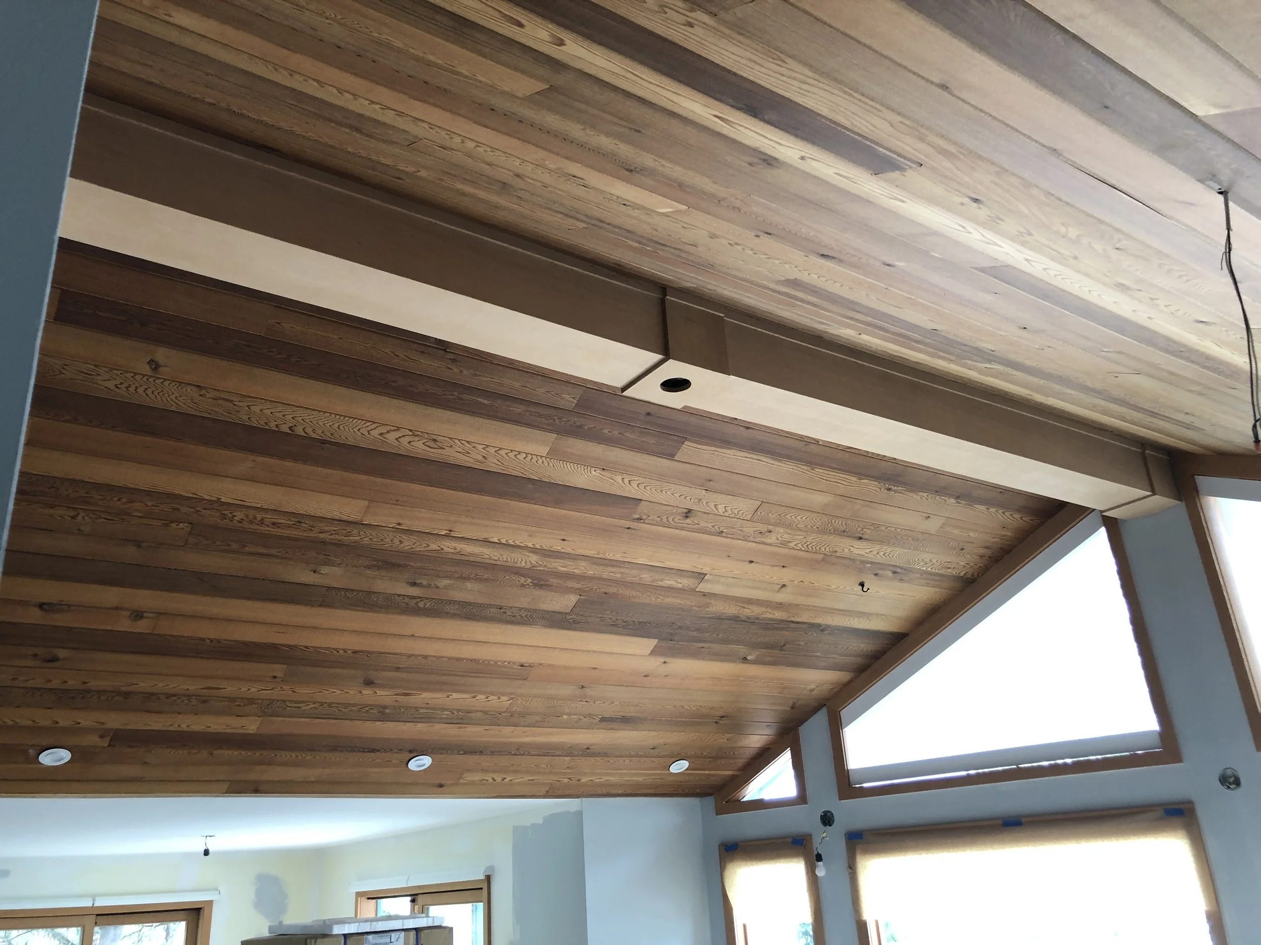 Interior ceiling with wooden planks, some recessed lighting, and a section of a light-colored wall with windows letting in natural light.
