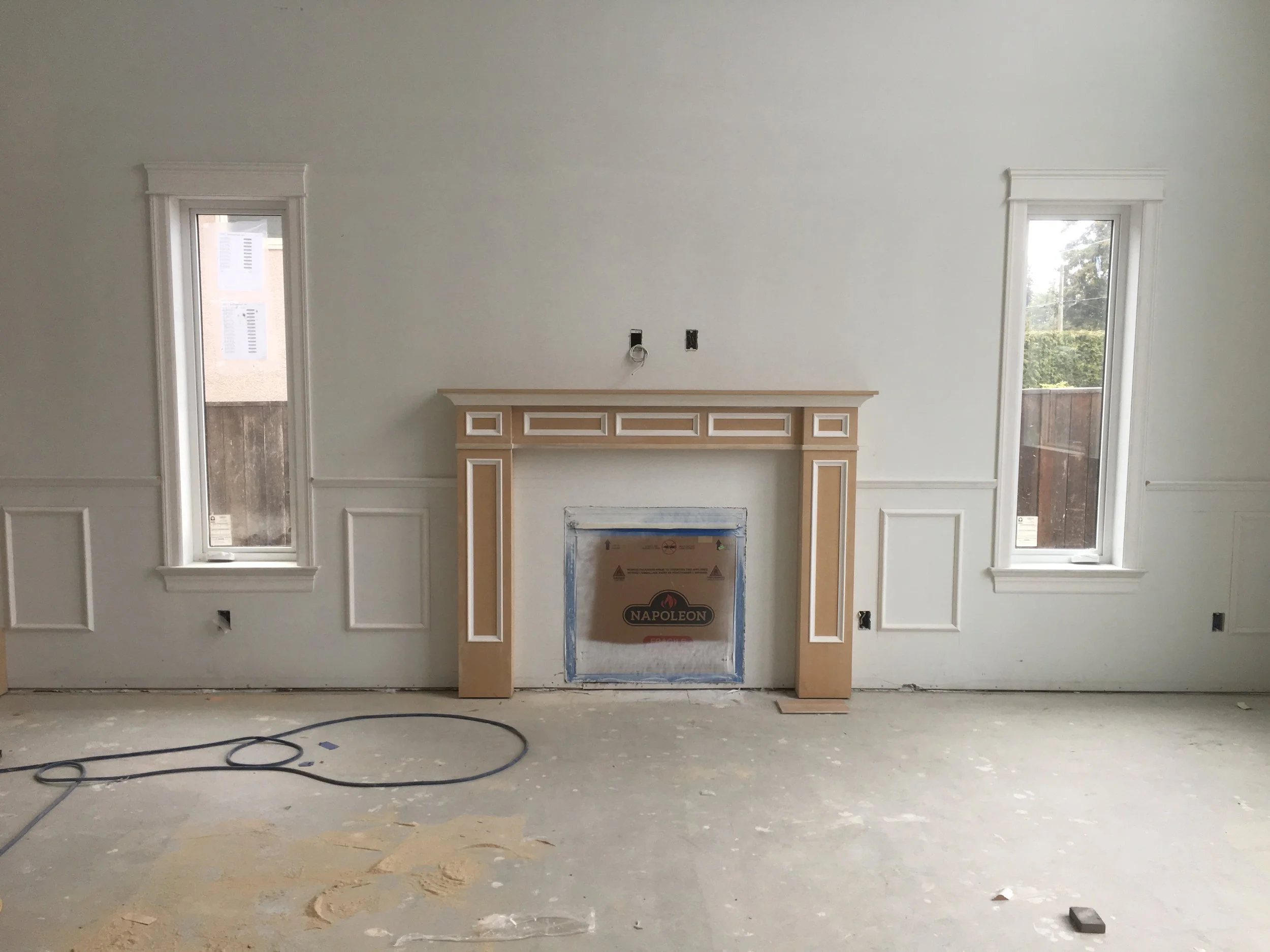 Interior room under construction with two small windows, a fireplace with decorative framing, and exposed wiring on the wall.