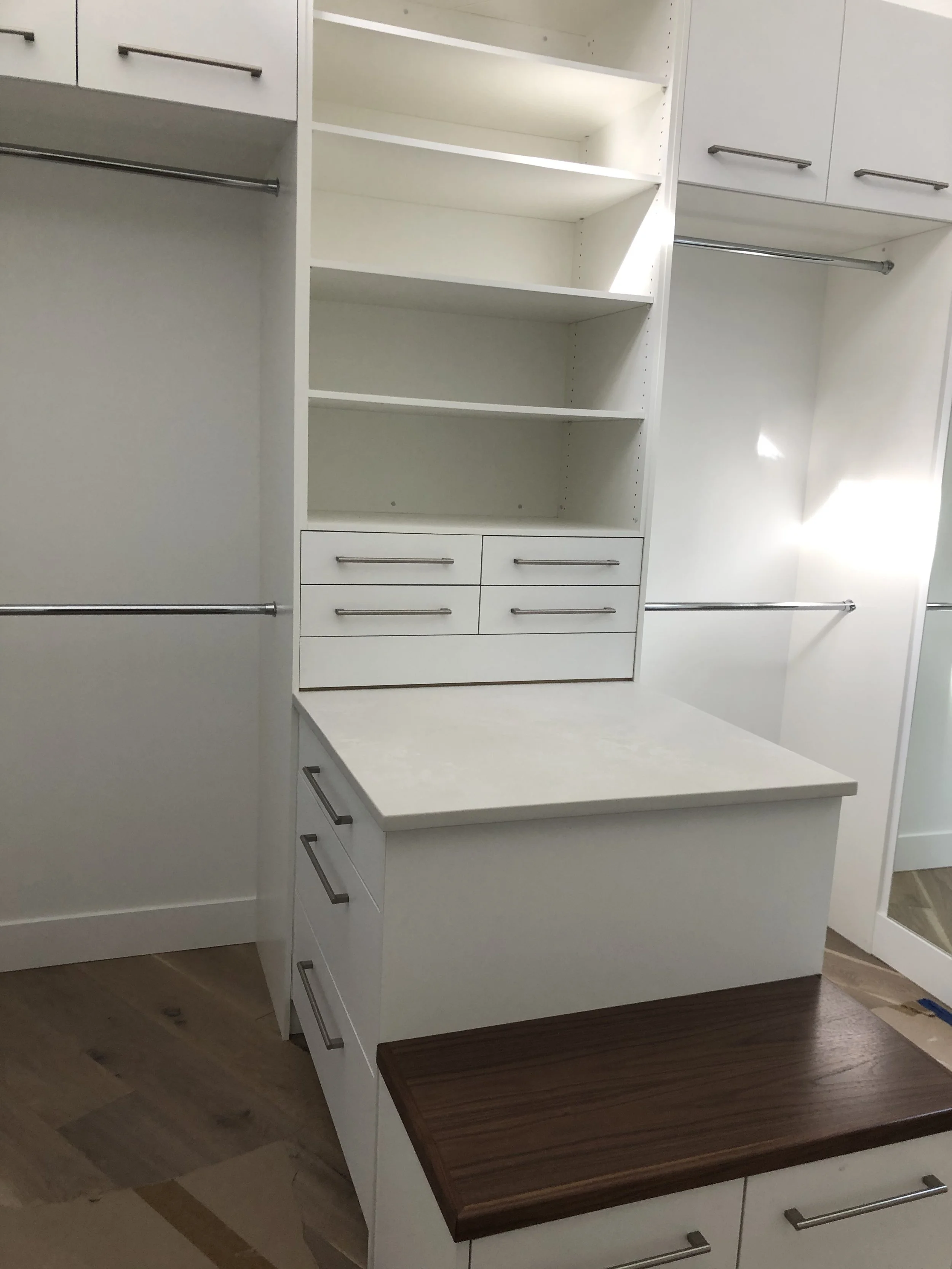 A white custom closet with shelves, drawers, and hanging rods. There is a dark wood countertop or bench at the bottom.