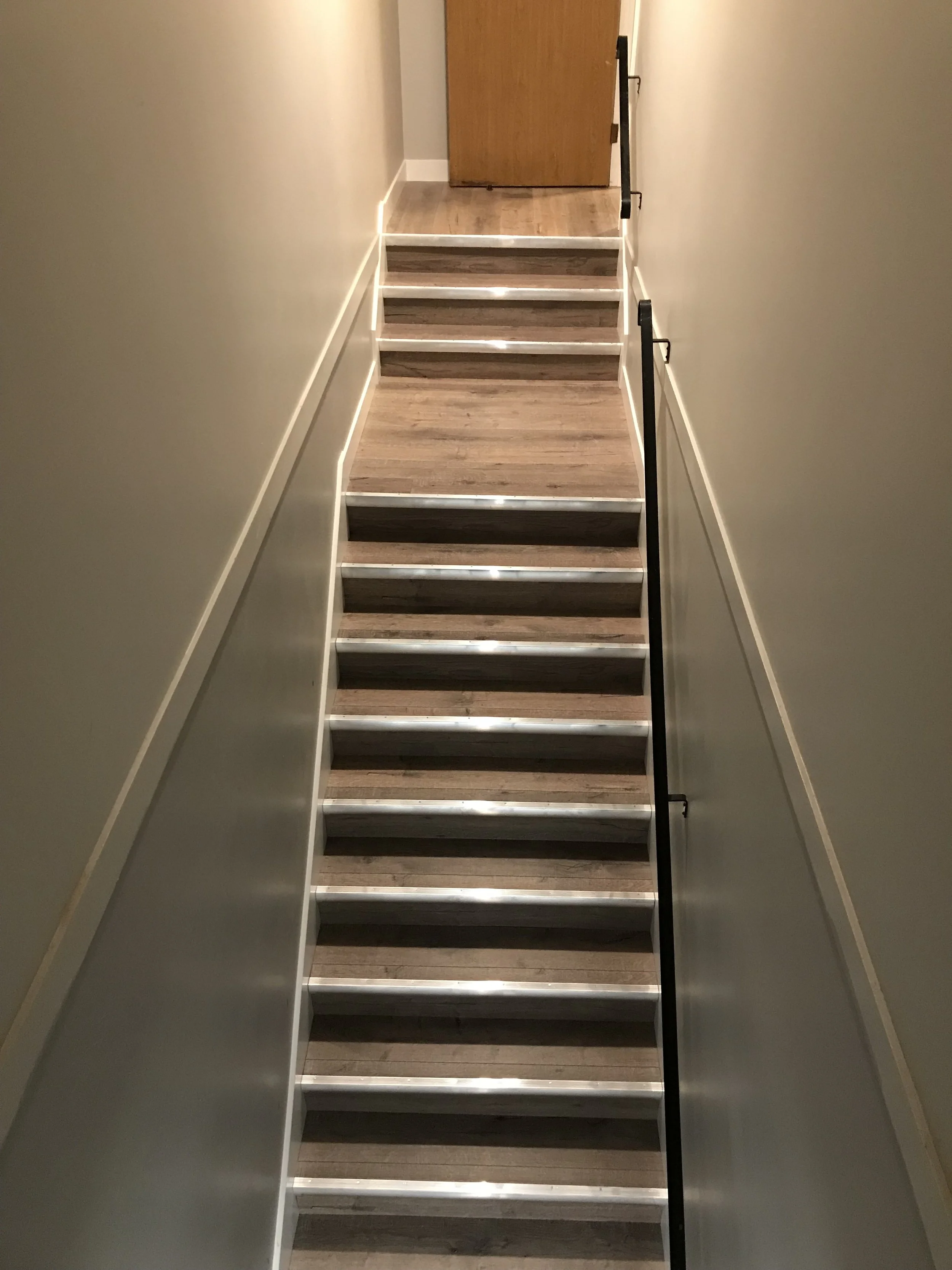 A narrow stairway with six steps leading to a closed wooden door at the top. The hallway walls are painted a light, neutral color, and there is a black handrail on the right side of the stairs.