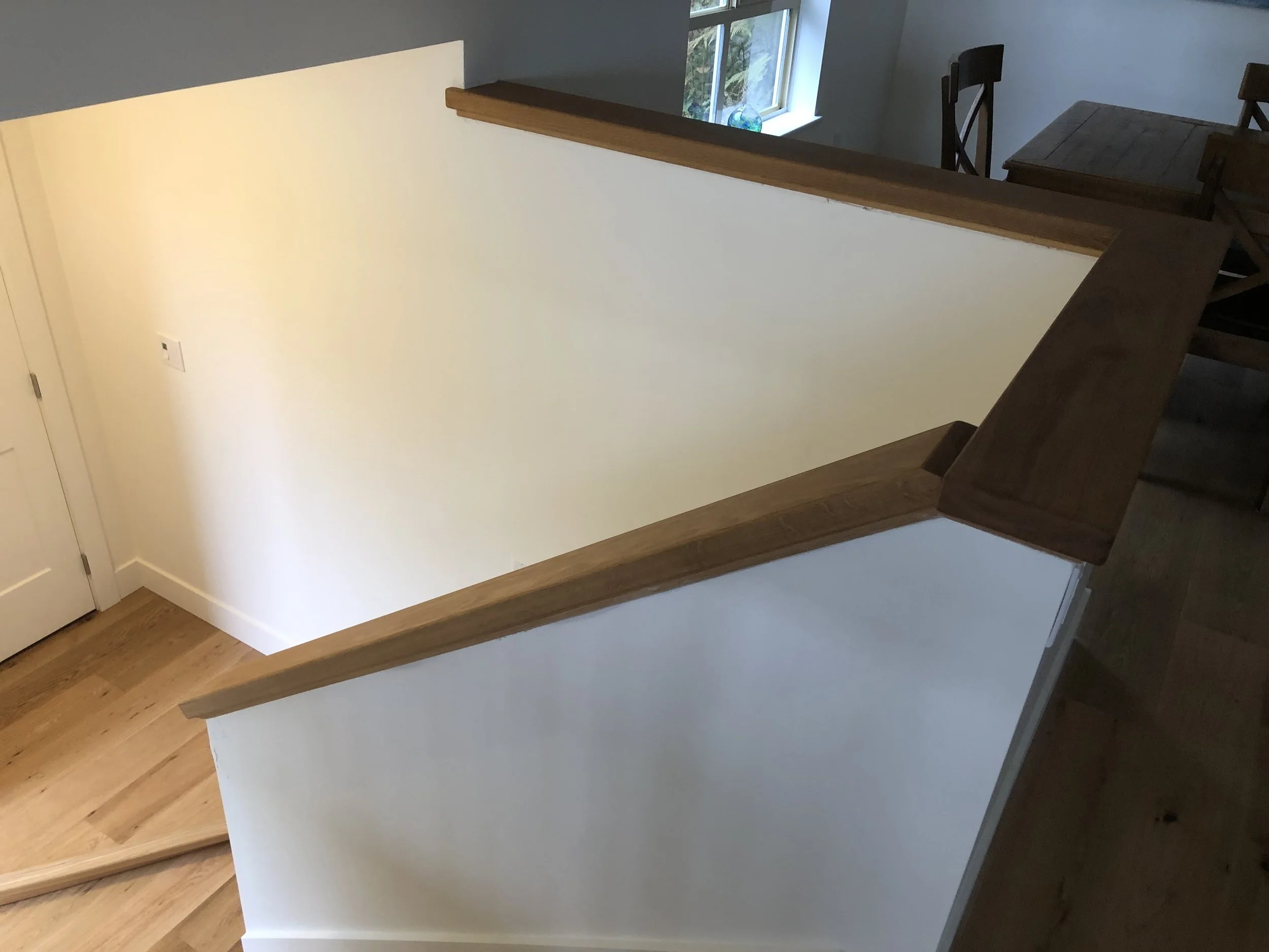 The image shows a staircase with wooden steps, a light-colored wall, and a section of a dining area with chairs and a table near a window.