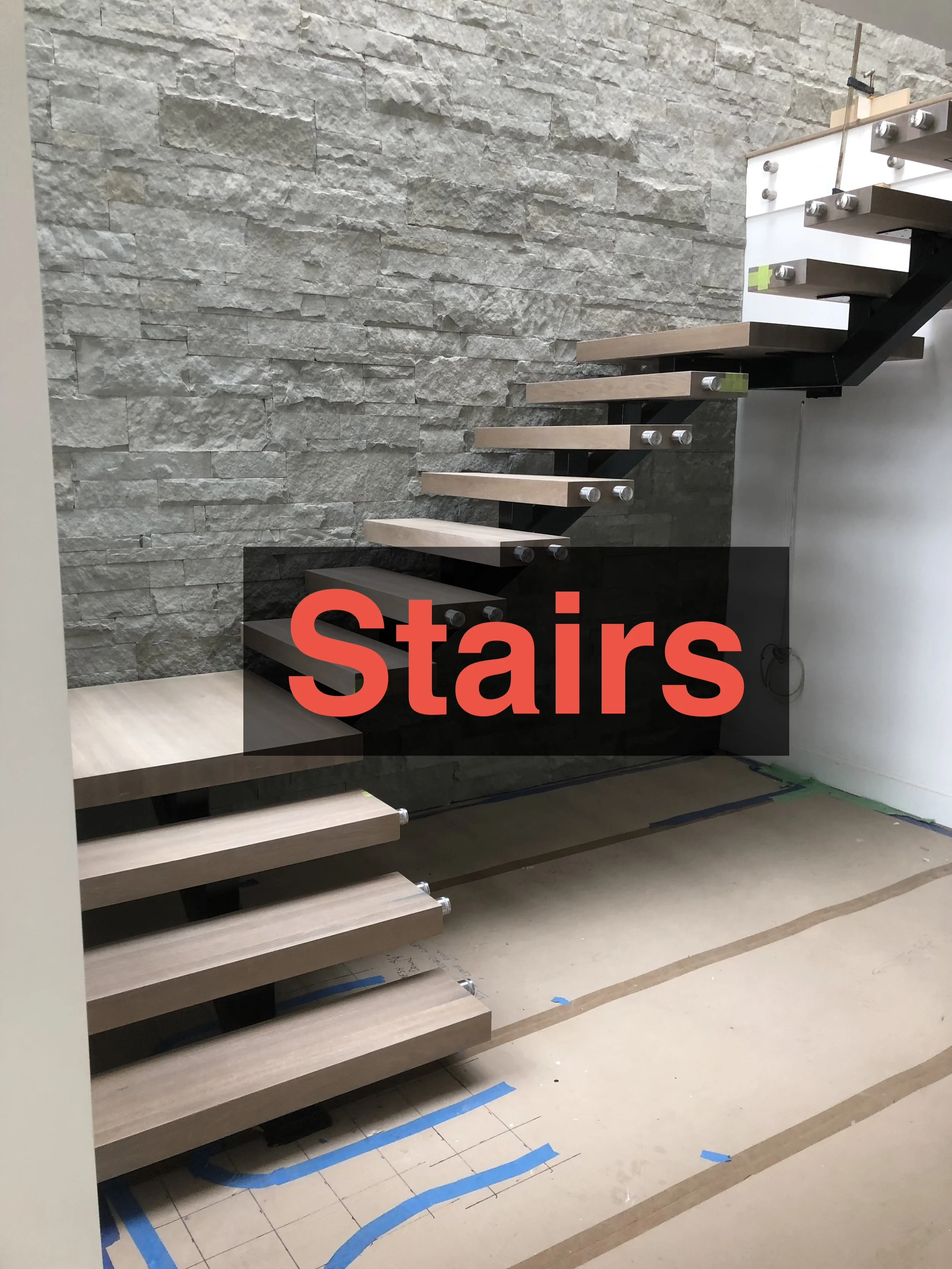 Unfinished staircase with light wood treads and dark metal supports, against a textured stone wall, with construction markings on the floor.