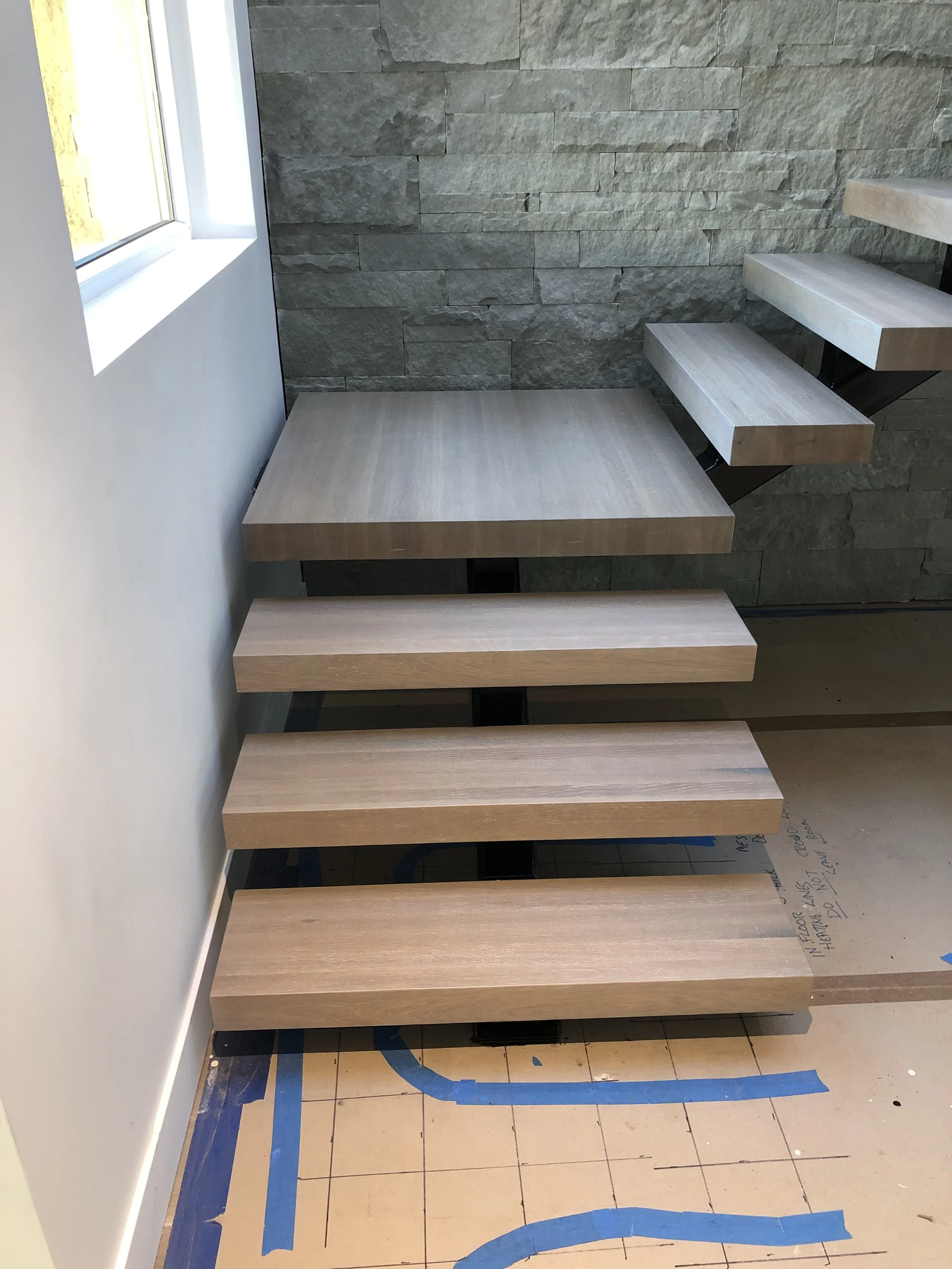 Interior of a room under construction with wooden stairs, a stone wall, a window, and blue painter's tape on the floor.
