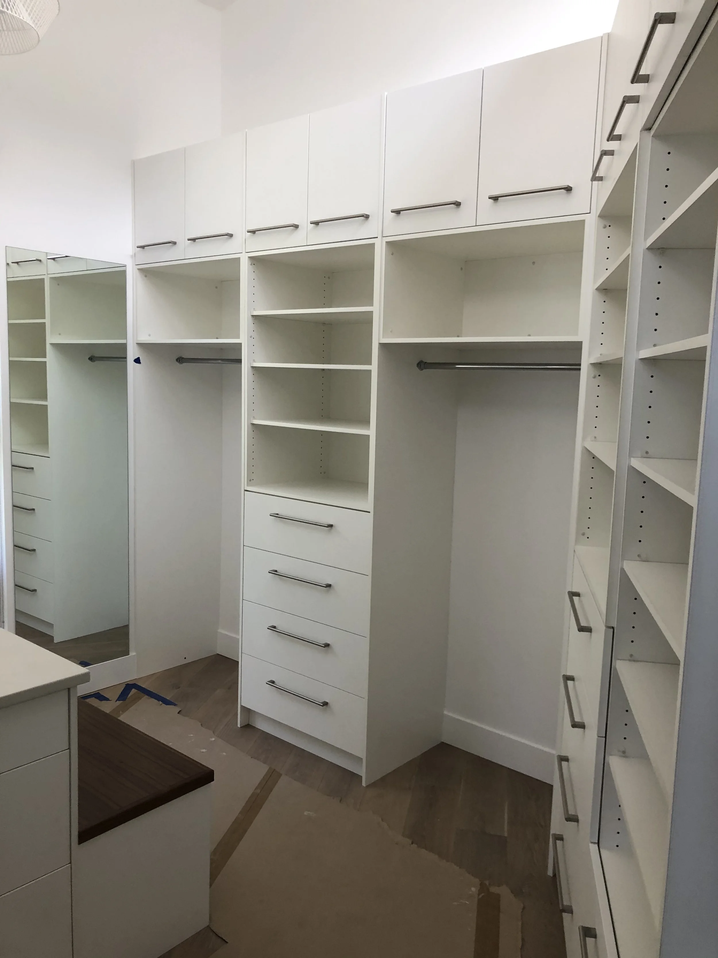 A room with white built-in shelves and cabinets, some with drawers and metal handles. There is a rectangular mirror and a wooden surface on a cabinet. The room appears to be in the process of being furnished or renovated.