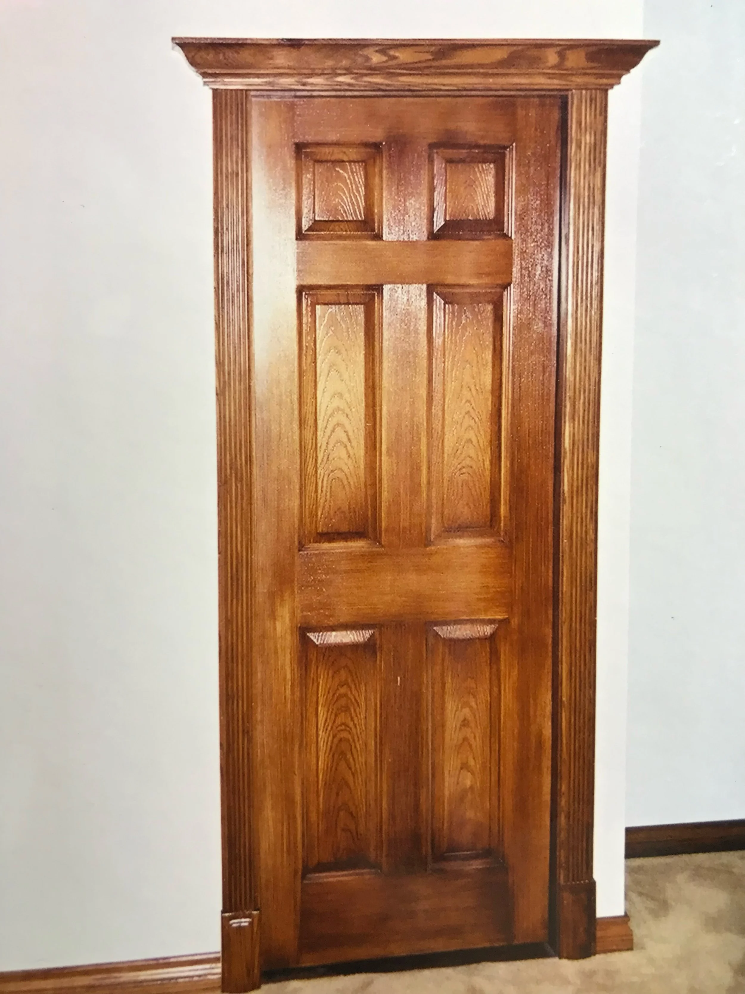 A tall wooden door with decorative raised panels and a detailed wooden frame. The door is closed and set against a plain white wall.