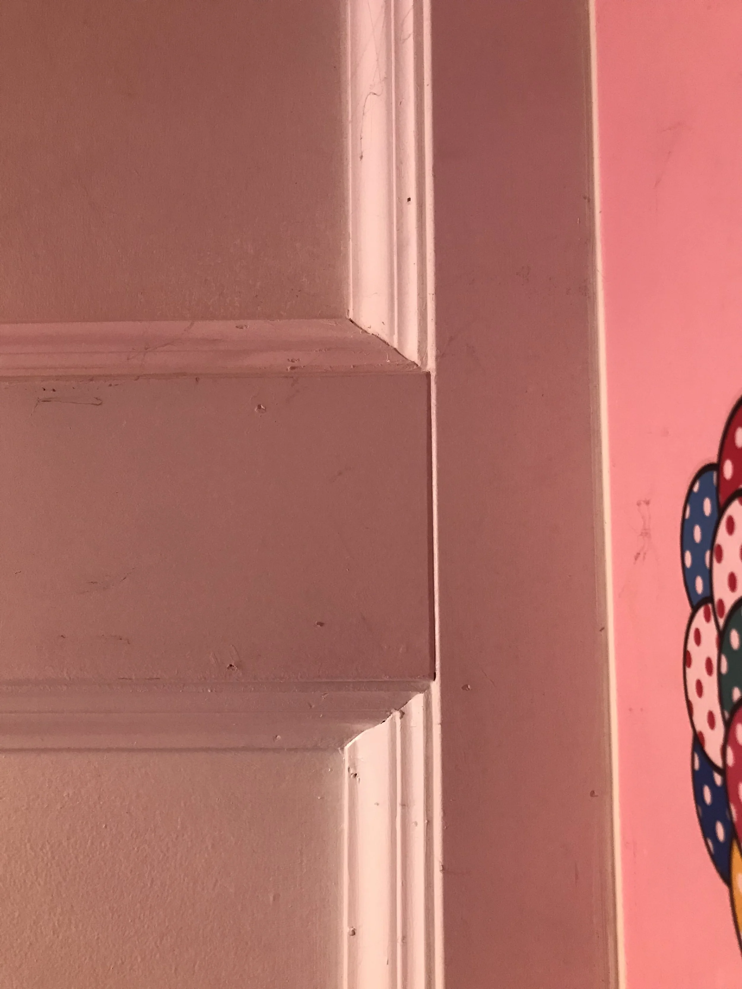 This is a close-up of a textured wall with a white baseboard at the bottom. The wall is painted in a shade of pink, with some minor imperfections and marks visible. On the right side of the image, there is a partial view of a colorful, patterned obje