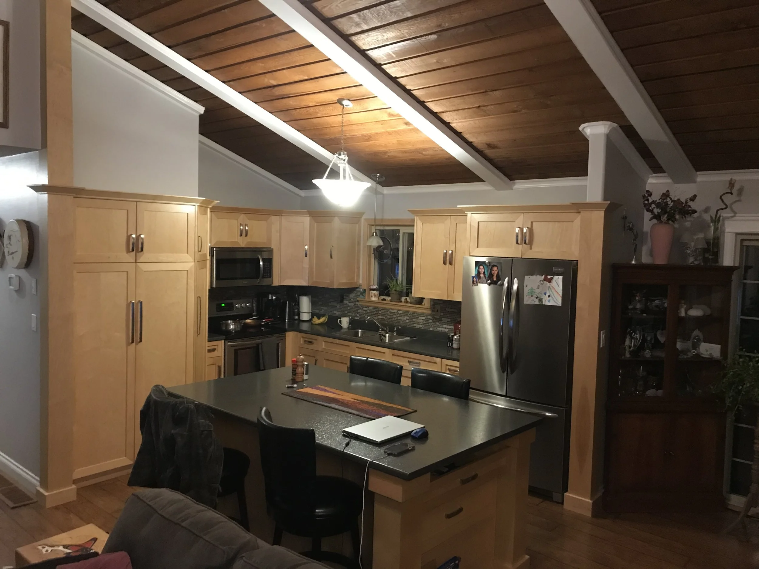 A kitchen with light wood cabinets, stainless steel refrigerator, and black countertops. The ceiling is wood-paneled with recessed lighting and a central chandelier. There is a dining table with chairs in the foreground, and various household items i