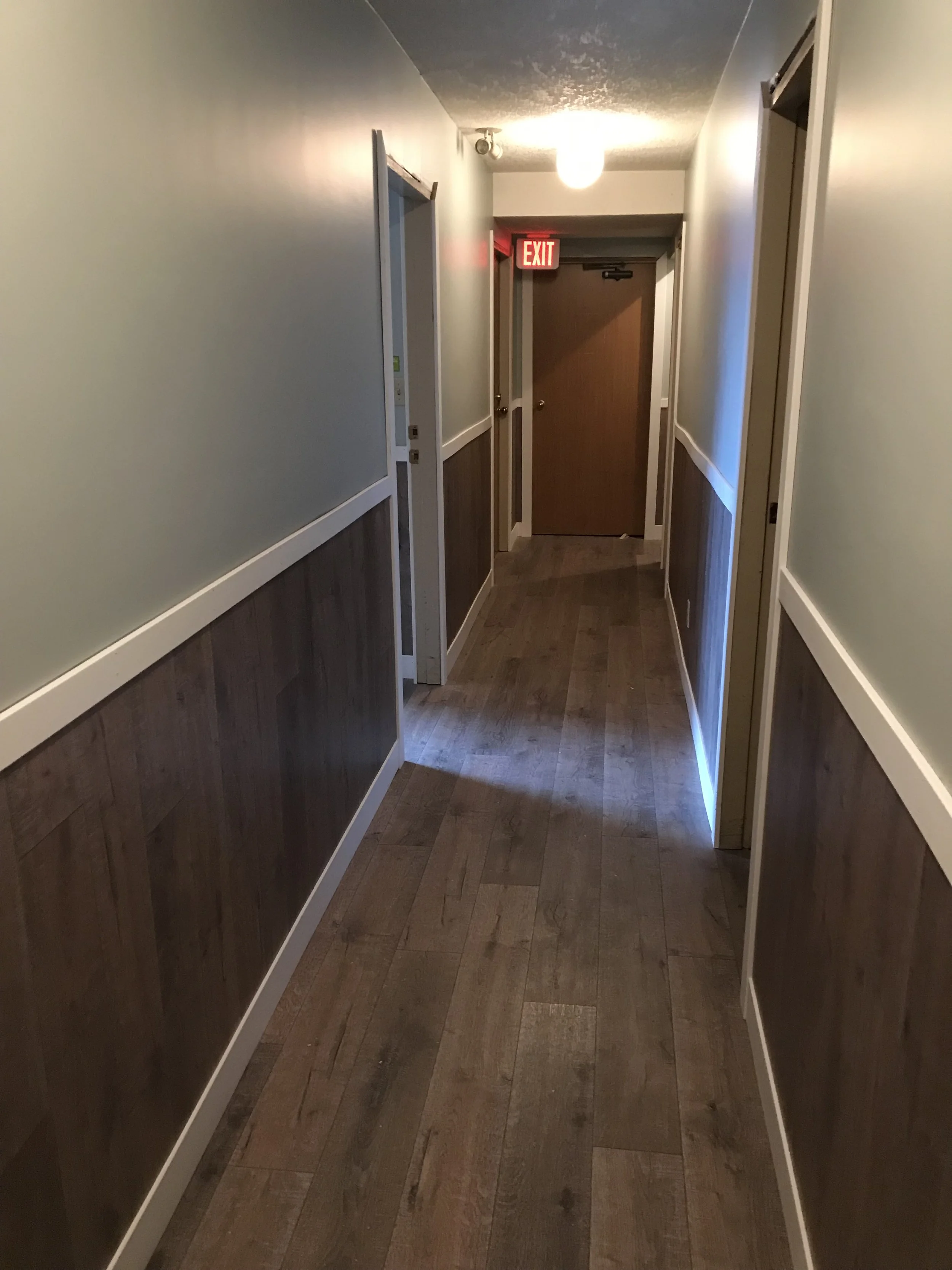 A hallway with wood paneled walls up to about waist height, painted light yellow above. White trim runs along the middle of the walls. The floor is wood, and there is a closed wooden door at the end of the hallway. Above the door is a red illuminated