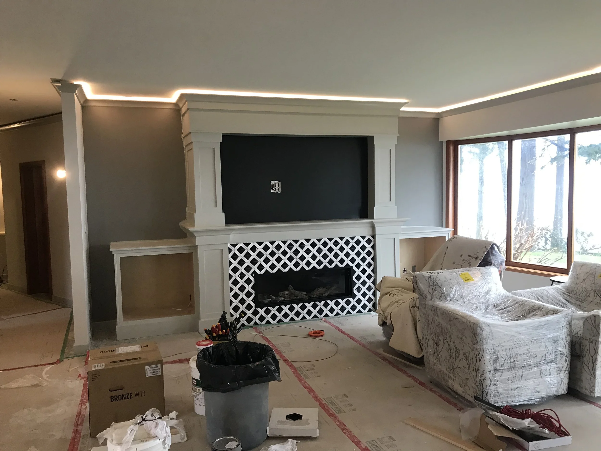 Living room under renovation with furniture covered, construction supplies, and tools scattered on the floor, an incomplete fireplace, and large windows showing trees outside.