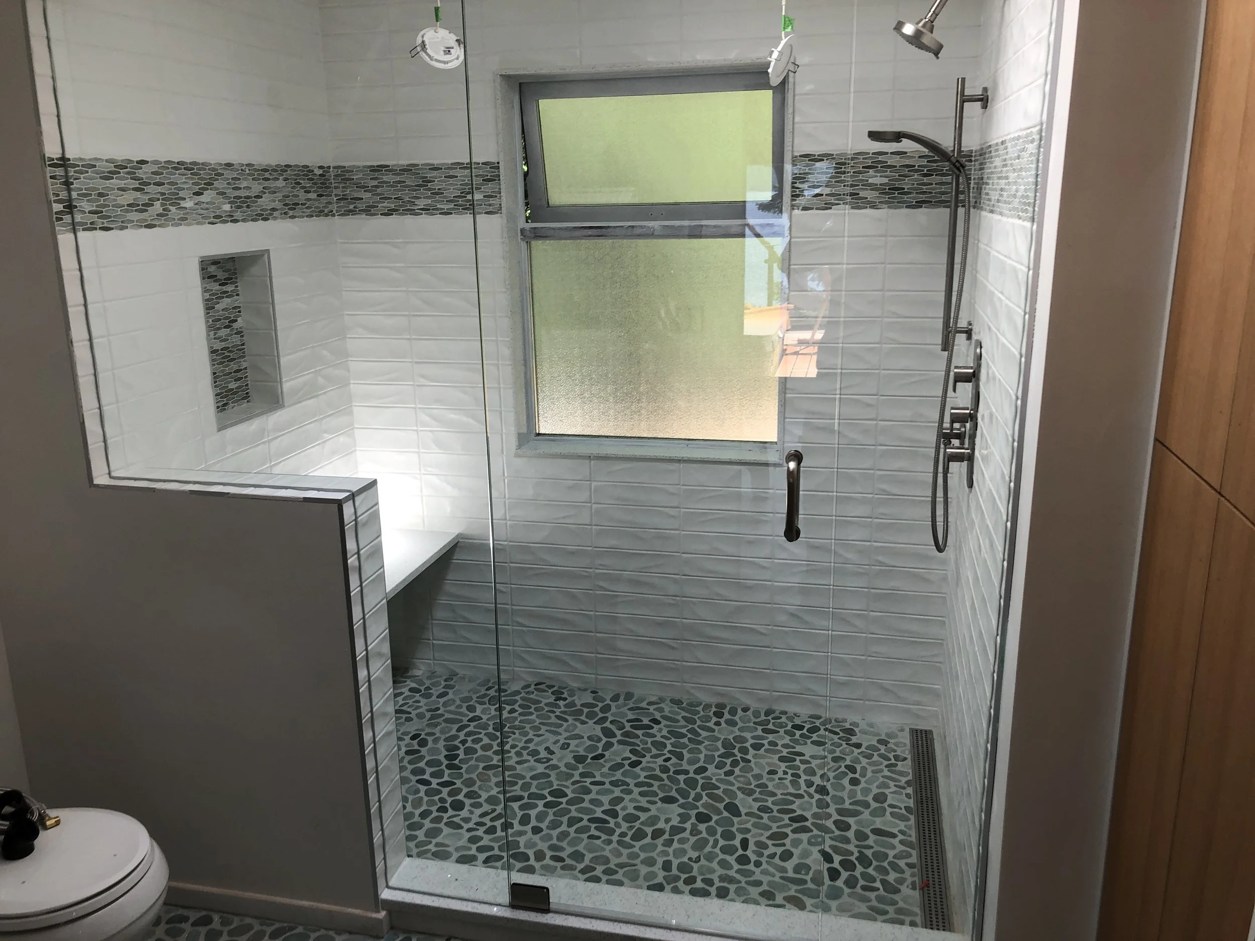Modern bathroom with a glass shower enclosure, white tiled walls featuring a decorative gray mosaic stripe, a niche in the shower wall, a small window, and a handheld showerhead, with wooden flooring outside the shower.