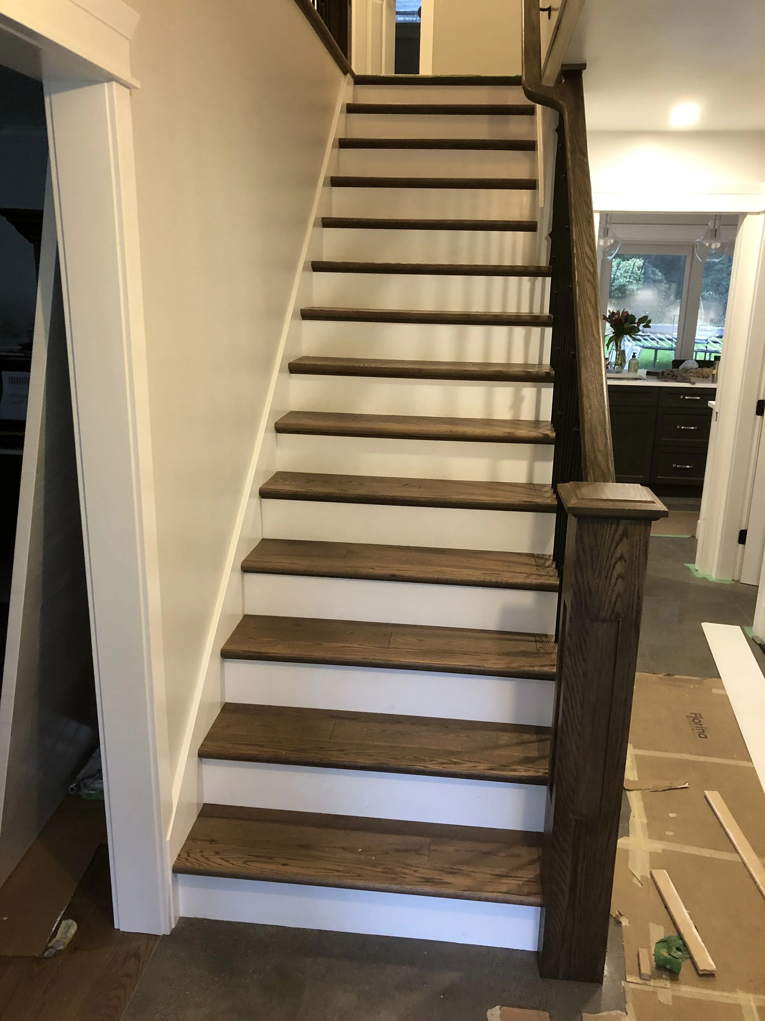 Indoor staircase with wooden steps and a dark wood handrail, leading to an upper floor. The staircase is next to a wall painted white, with a kitchen visible in the background through a doorway, featuring a window with flowers on the counter.