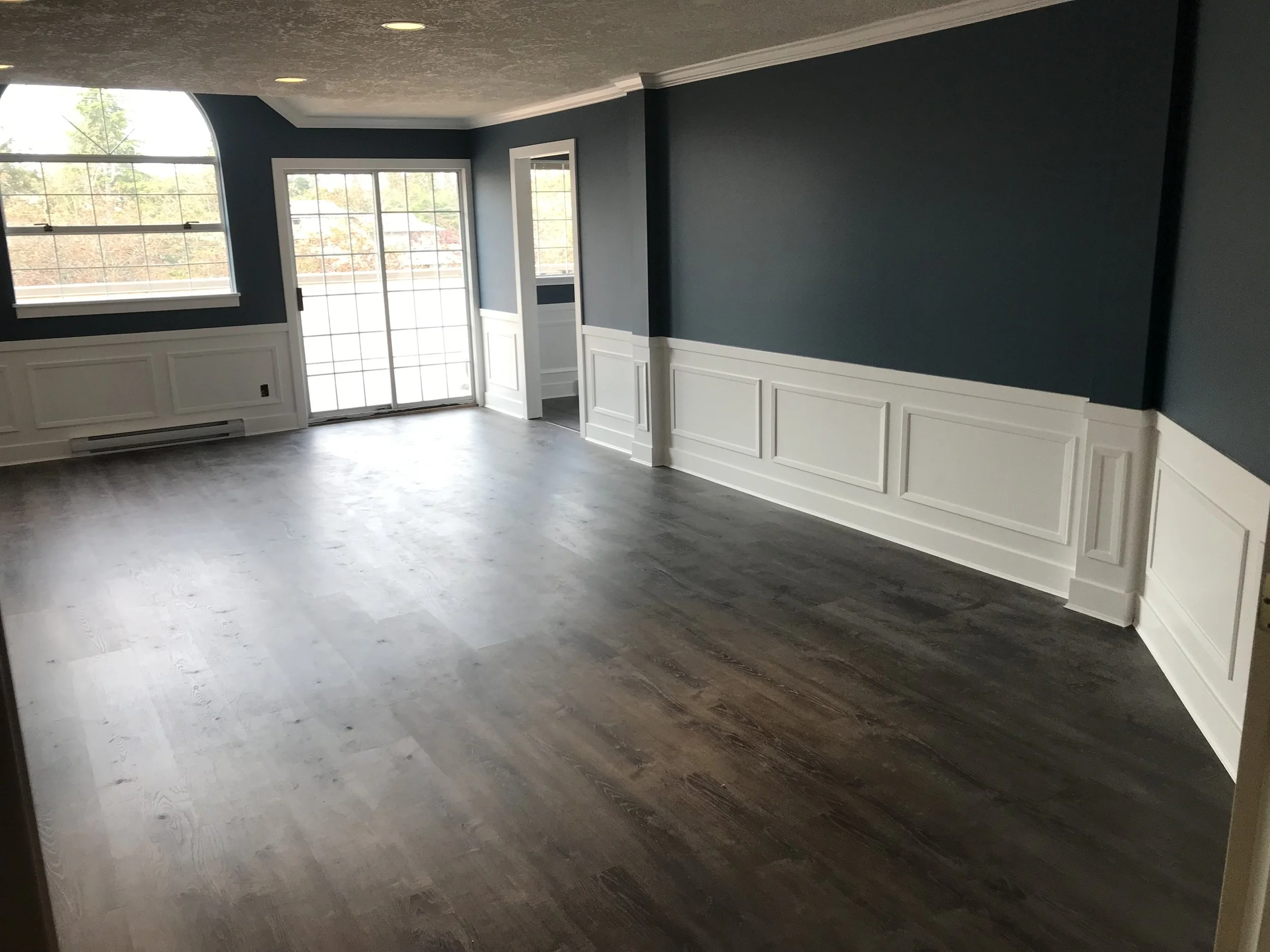 Empty room with dark wood flooring, blue walls with white paneling, large windows, and a sliding glass door leading outside.