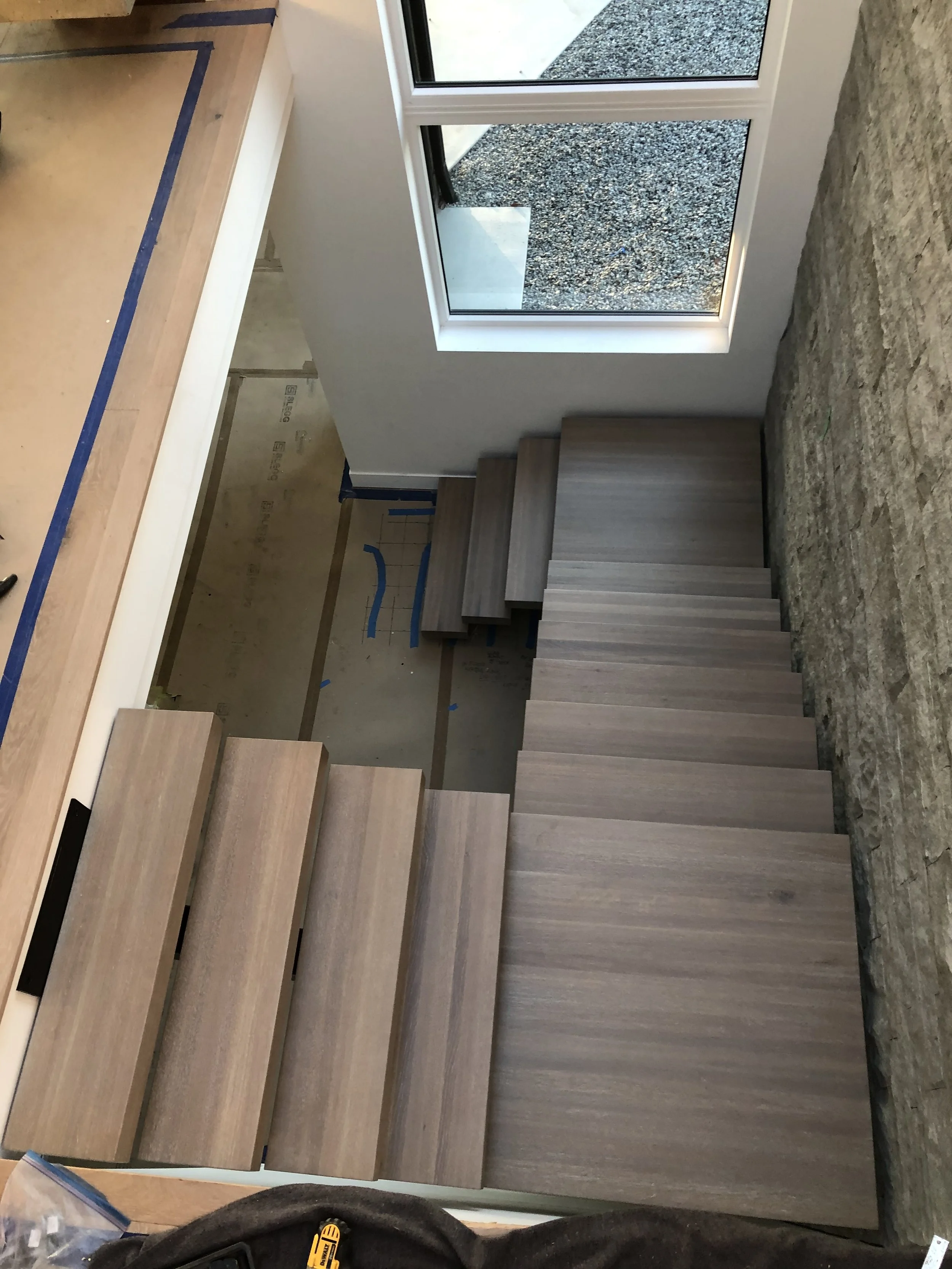 Interior of a room under construction with wooden stairs leading to an upper level, a large window showing an outdoor gravel area, and unfinished wall space with visible insulation and wiring.