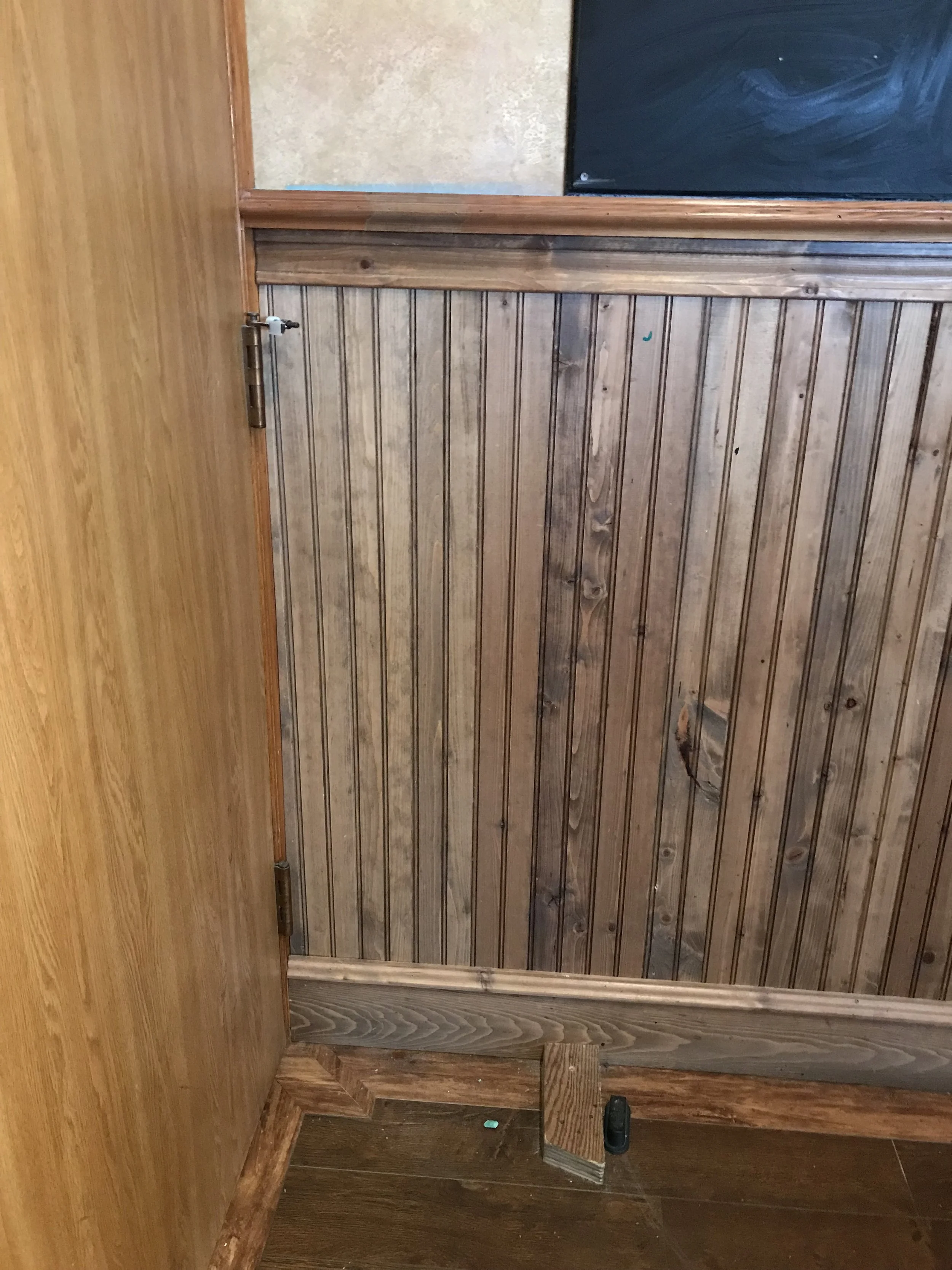 A wooden pet gate with a latch, installed in a room with wood paneling on the lower half of the wall and a black chalkboard on the upper part. The floor is also wooden.