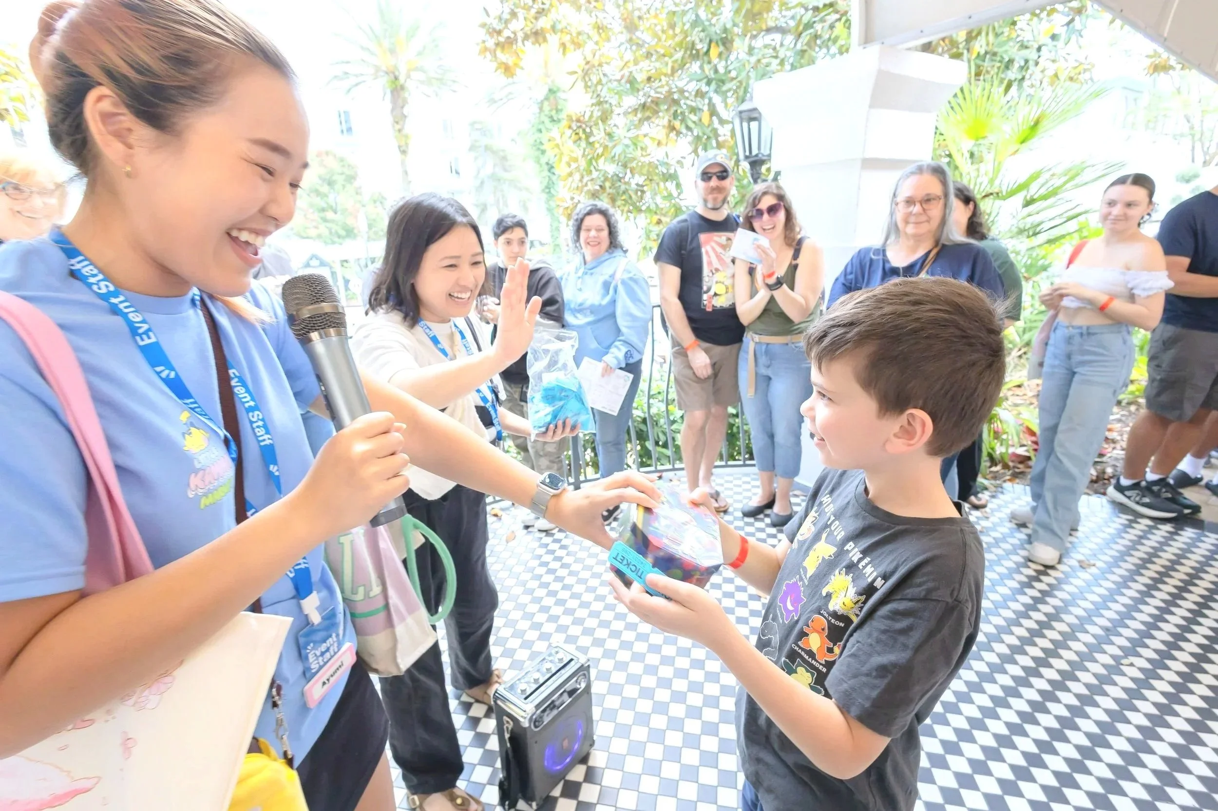 A group of people gathered outdoors, with some smiling and watching as a woman hands a gift to a young boy. The woman is holding a microphone and wearing a blue shirt with event staff lanyard. The background shows trees and a sunny day.