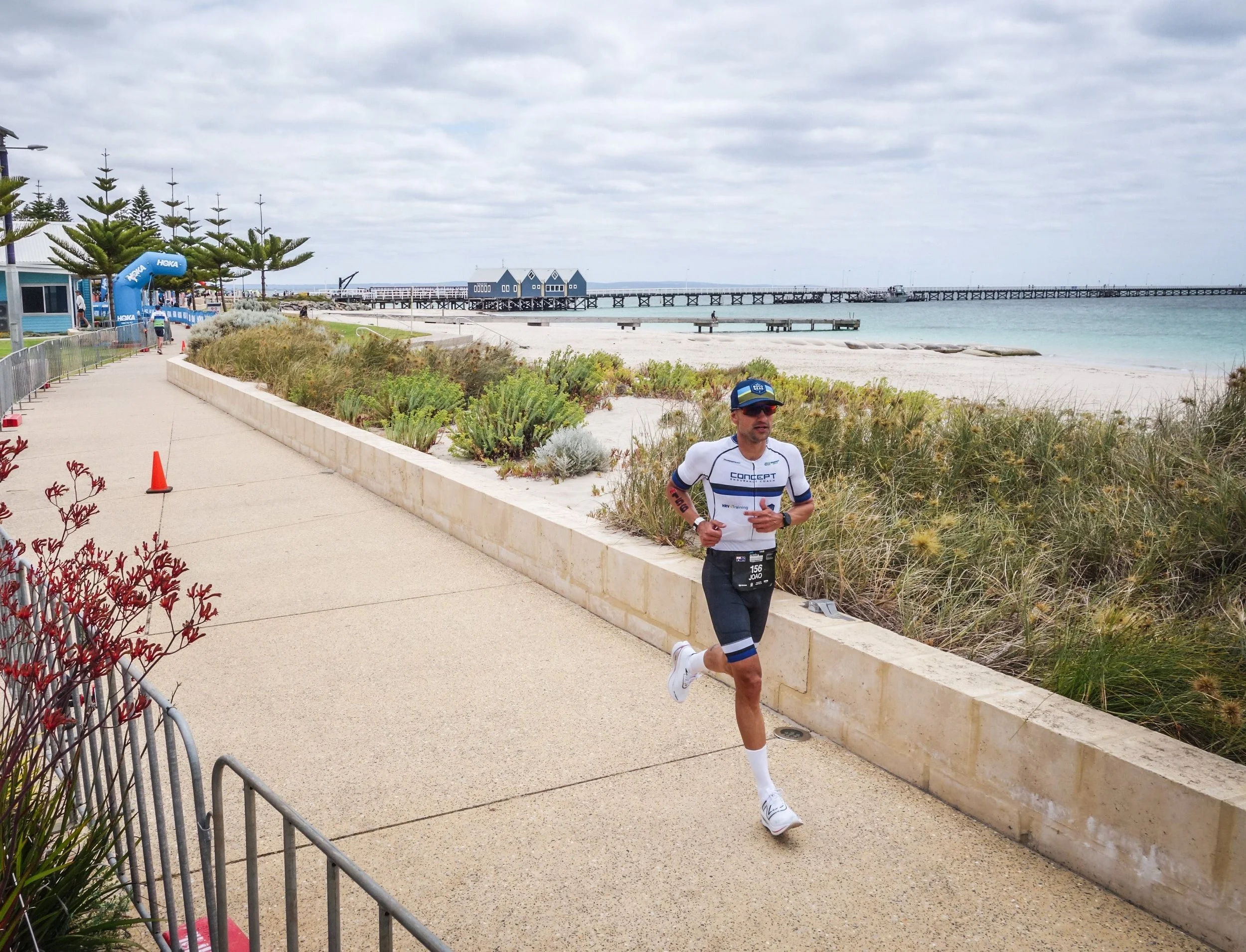 A man running along a paved pathway near a beach with a pier and a small blue building in the background, under a cloudy sky.