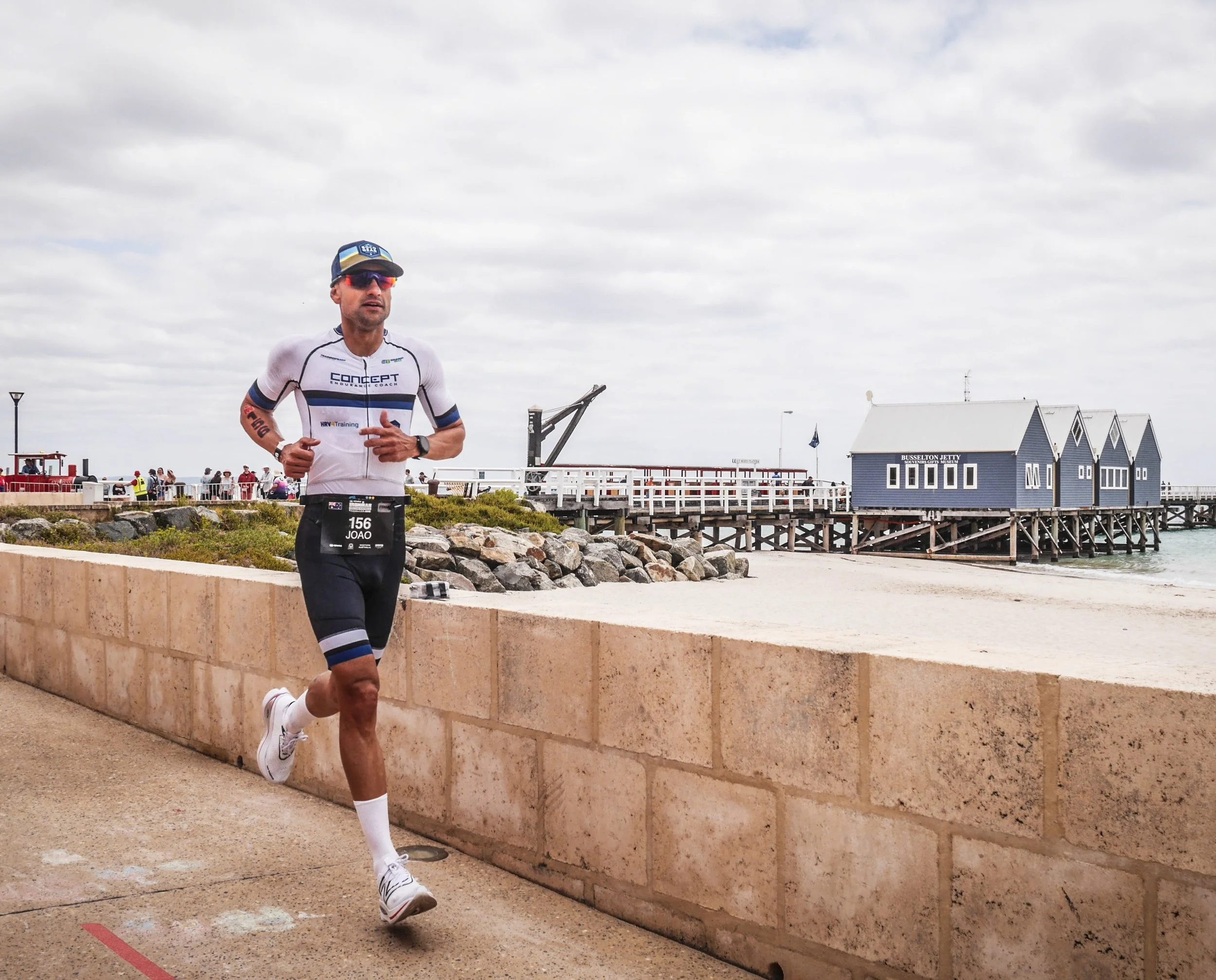 A male runner in athletic gear with a race bib running along a seaside pathway near buildings and a pier, with cloudy skies overhead.