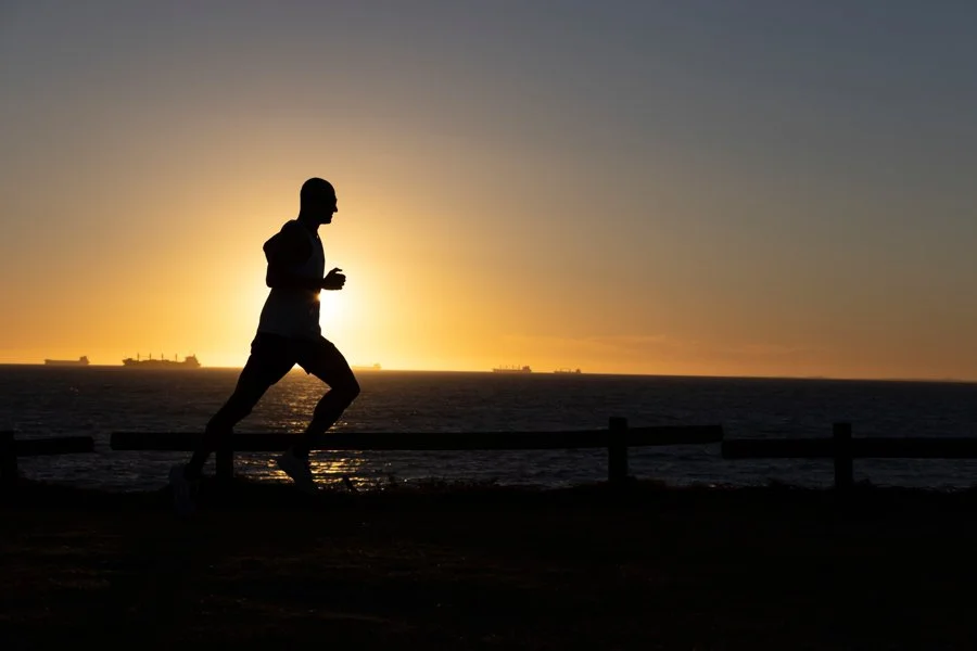 Silhouette of a person jogging along the coast during sunset, with ships visible on the horizon.