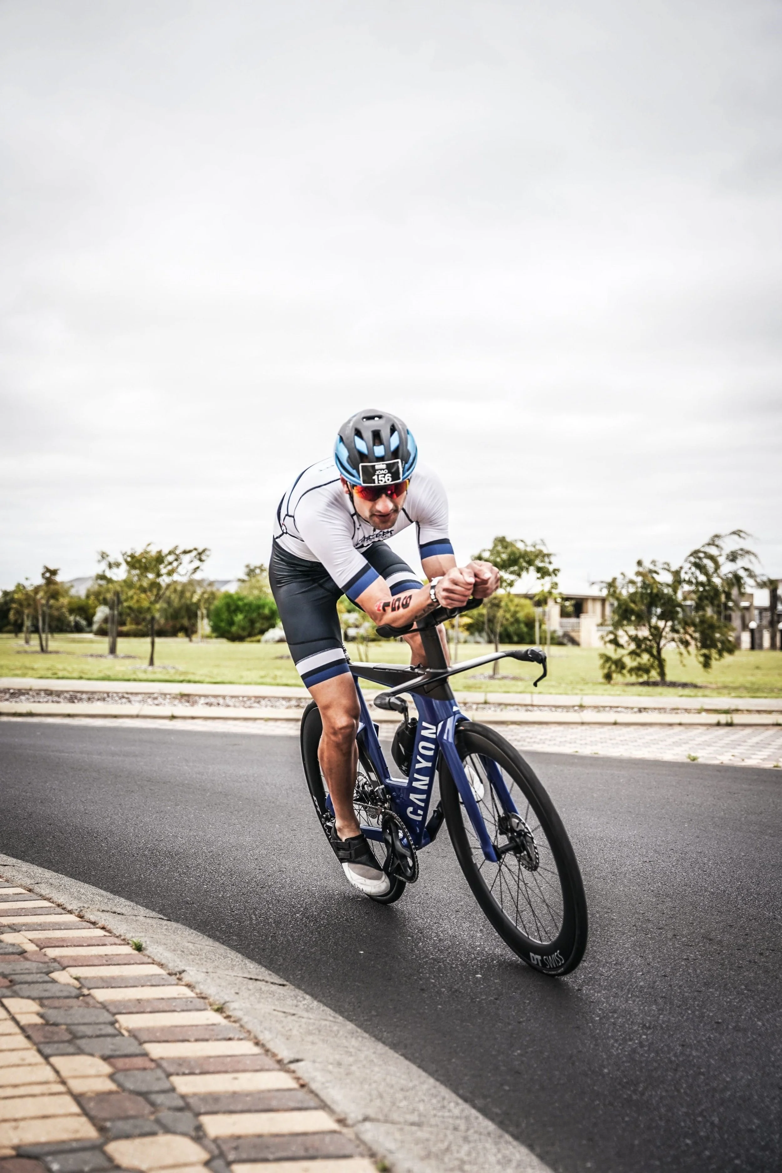 Cyclist in white and blue athletic clothing and helmet leaning into a turn on a black road bike with a city park background under an overcast sky.