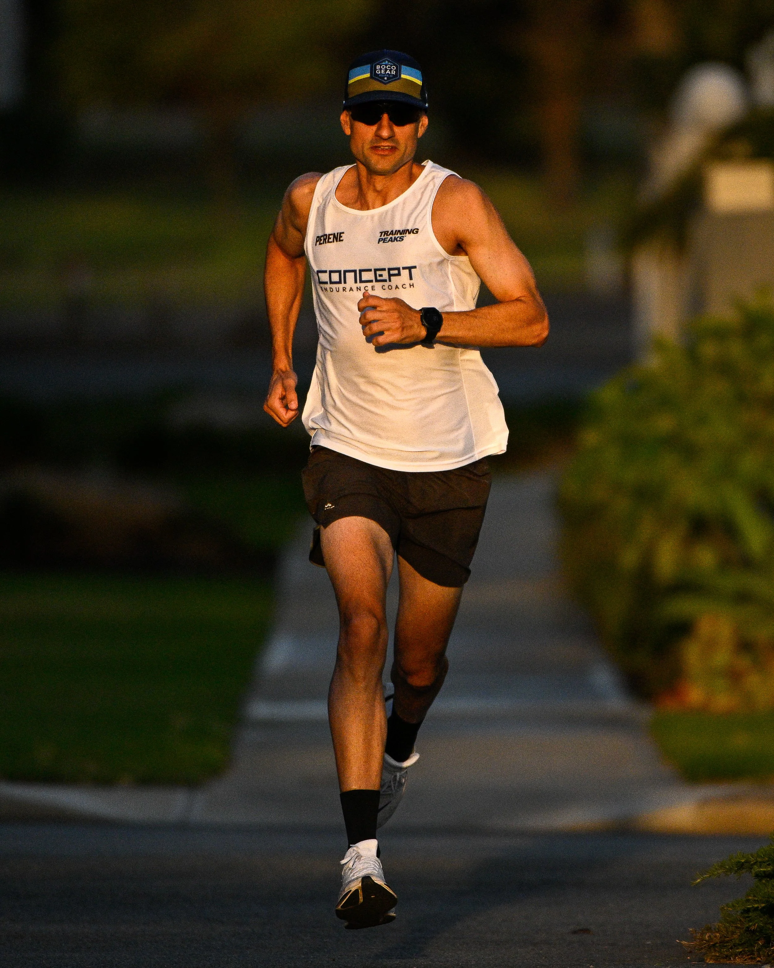 A man running outdoors during the evening or early morning, wearing a white sleeveless athletic shirt, black shorts, a blue cap, sunglasses, and a fitness watch.