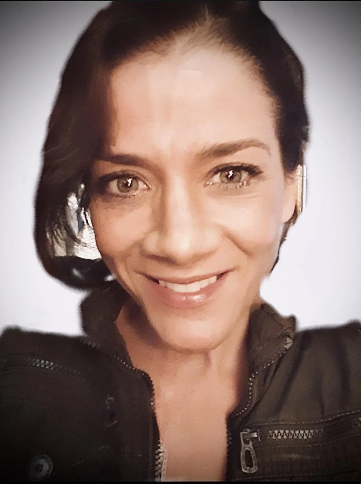 A woman with short dark hair and hazel eyes smiling at the camera, wearing a black jacket with a zipper.