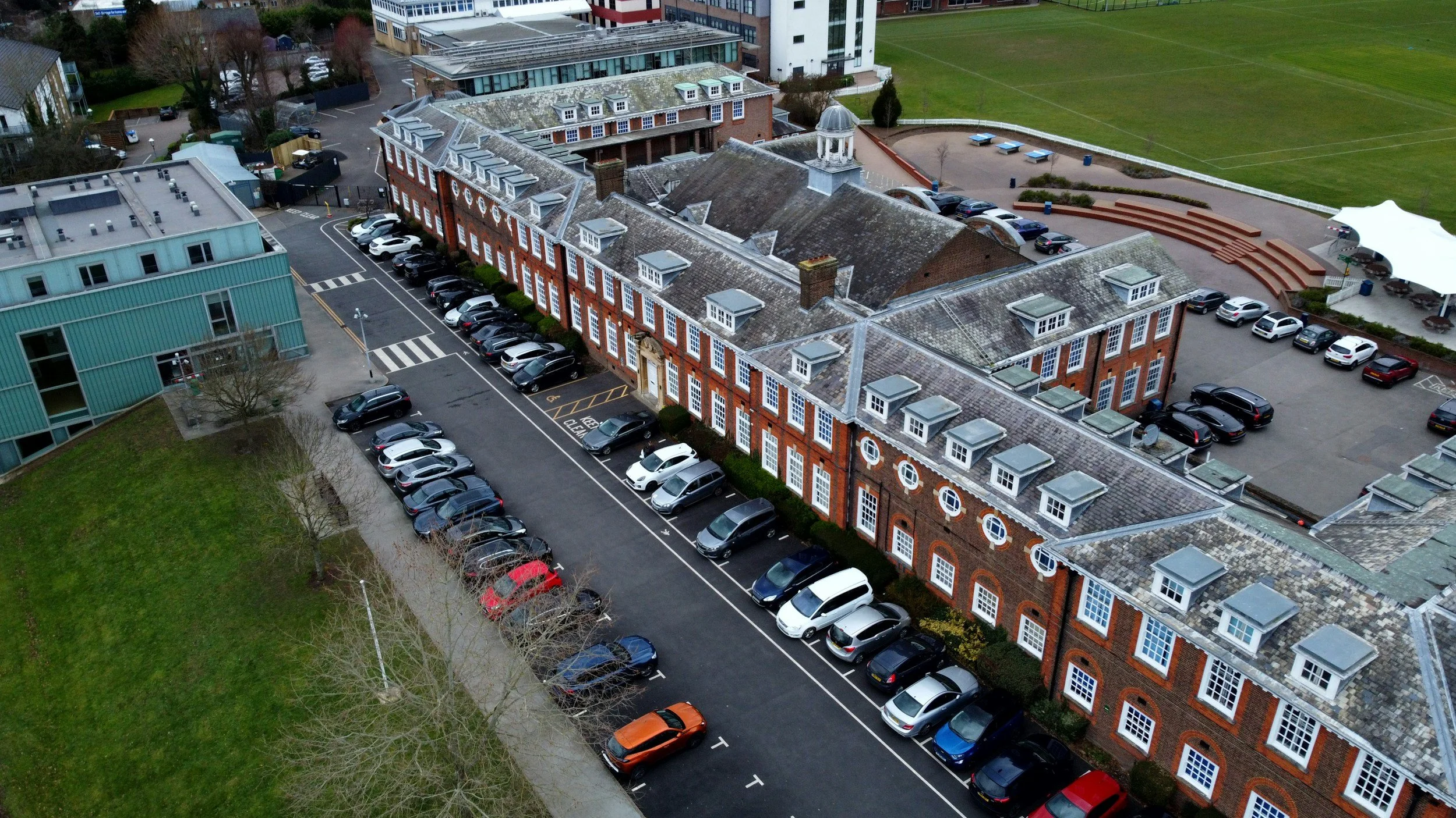 An aerial view of a red brick building with a gray slate roof and white window frames, adjacent to a parking lot with numerous cars, and a grassy sports field in the background.