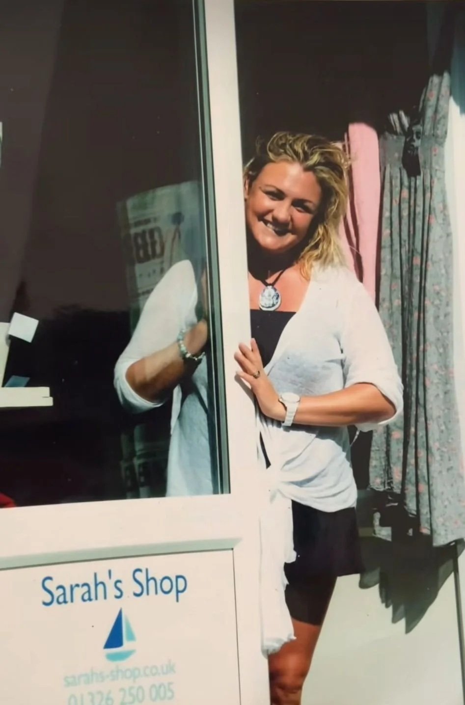 A woman with blonde hair smiling and peeking out from behind a glass door in front of a shop sign that reads "Sarah's Shop" with a website address and phone number underneath.