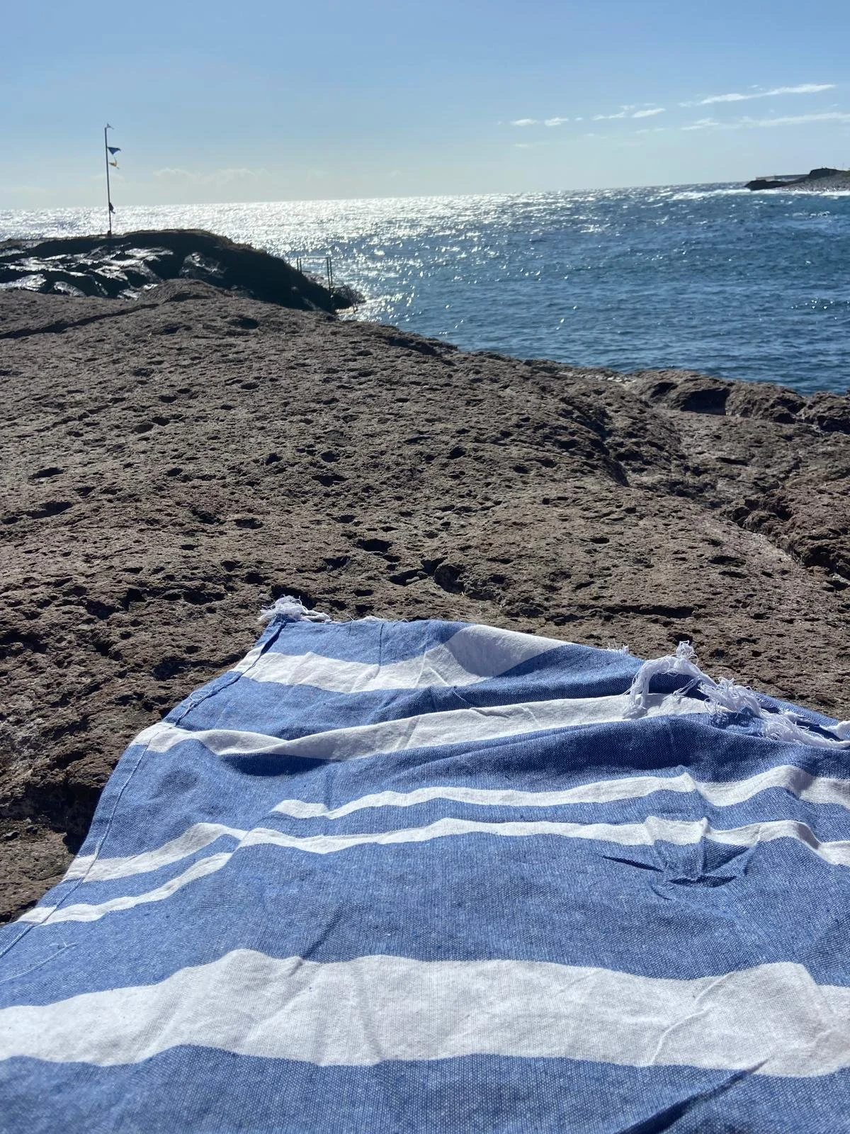 Blue and white striped towel on a rocky surface by the ocean, with the sea and a partly cloudy sky in the background.