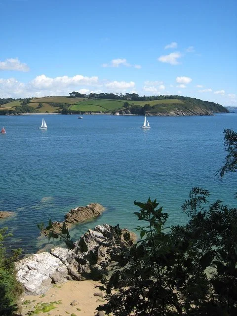 Coastal scene with blue water, sailboats, rocky shoreline, and green hills in the background under a partly cloudy sky.