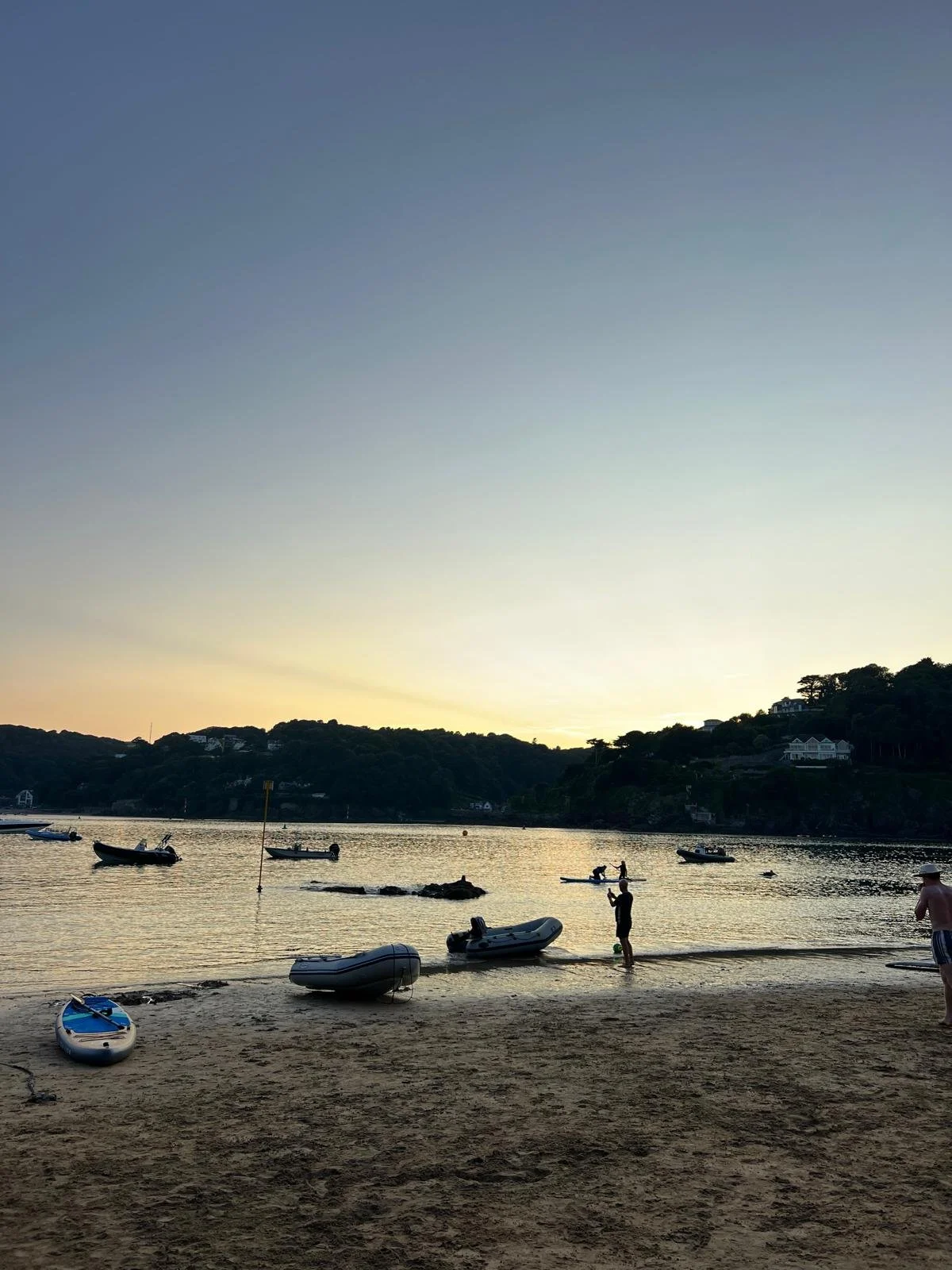 A peaceful lakeside scene at sunset with small boats floating on the water, a few people standing and enjoying the view, and houses on the hill in the background.