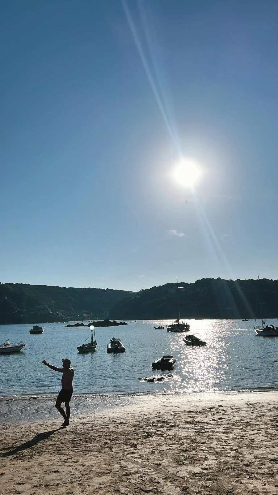 A person standing on a sandy beach with arms outstretched, facing calm water with sailboats and motorboats anchored, under a bright sun in a clear blue sky, with hills in the background.