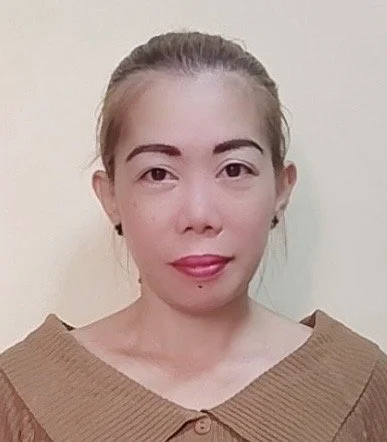 A woman with light skin, blonde hair tied back, wearing a tan sweater with a wide neckline, standing against a plain, light-colored wall.