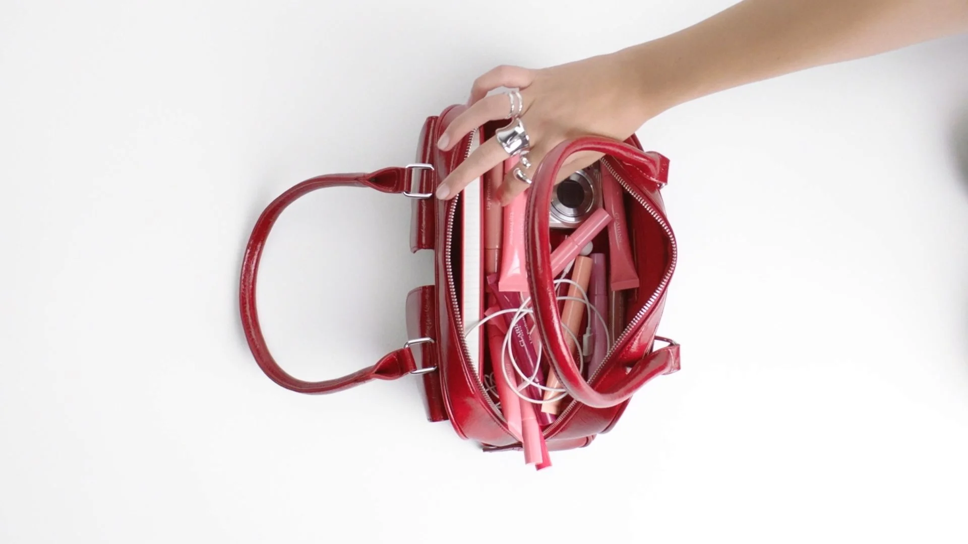 Open red handbag filled with pink and nude makeup items, brushes, and accessories on white background.
