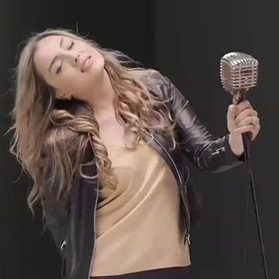 A young woman with long wavy hair singing with eyes closed, holding a vintage microphone, wearing a black leather jacket and a beige top, against a black background.