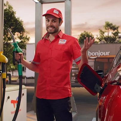 A gas station attendant wearing a red uniform and cap, holding a fuel nozzle, standing beside a red car at a fuel pump, with a Shell station and a Towne Mart sign in the background.