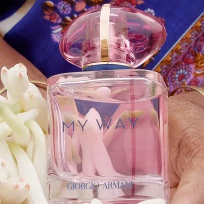 A pink Giorgio Armani perfume bottle with a gold and pink cap, held in someone's hand. There are flower petals nearby, and a blue and multicolored fabric in the background.