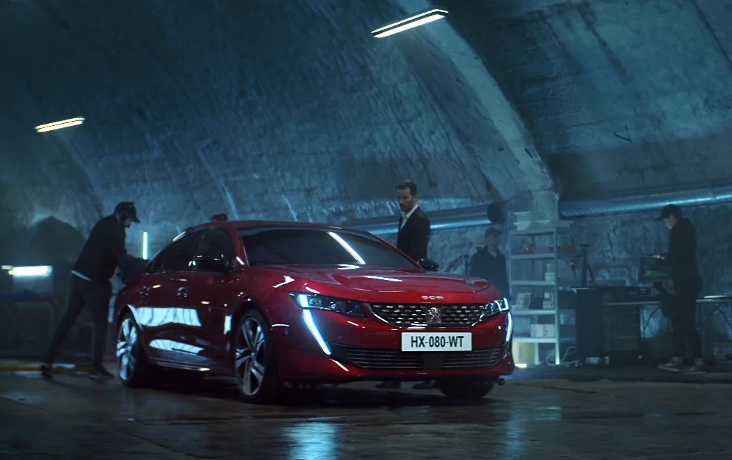 Red Peugeot sedan in a dimly lit, industrial-style garage with four people around it, some working on or inspecting the vehicle.