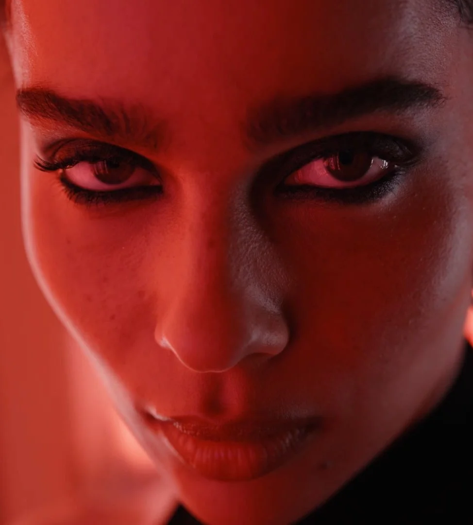 Close-up of a woman's face with intense expression, glowing skin, dark eyebrows and makeup, and reddish lighting.