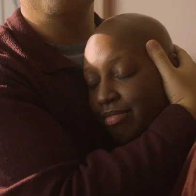 Close-up of a man gently holding and comforting a bald woman with closed eyes.