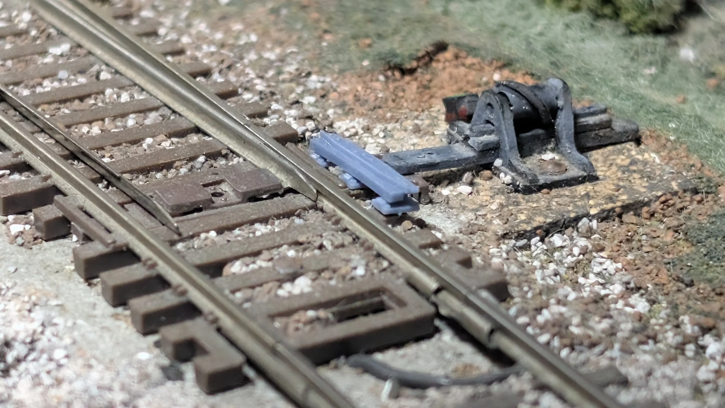 Close-up of railway tracks with a mechanically attached small blue device at the track joint and a black component on the ground nearby, with grass and gravel surrounding the tracks.