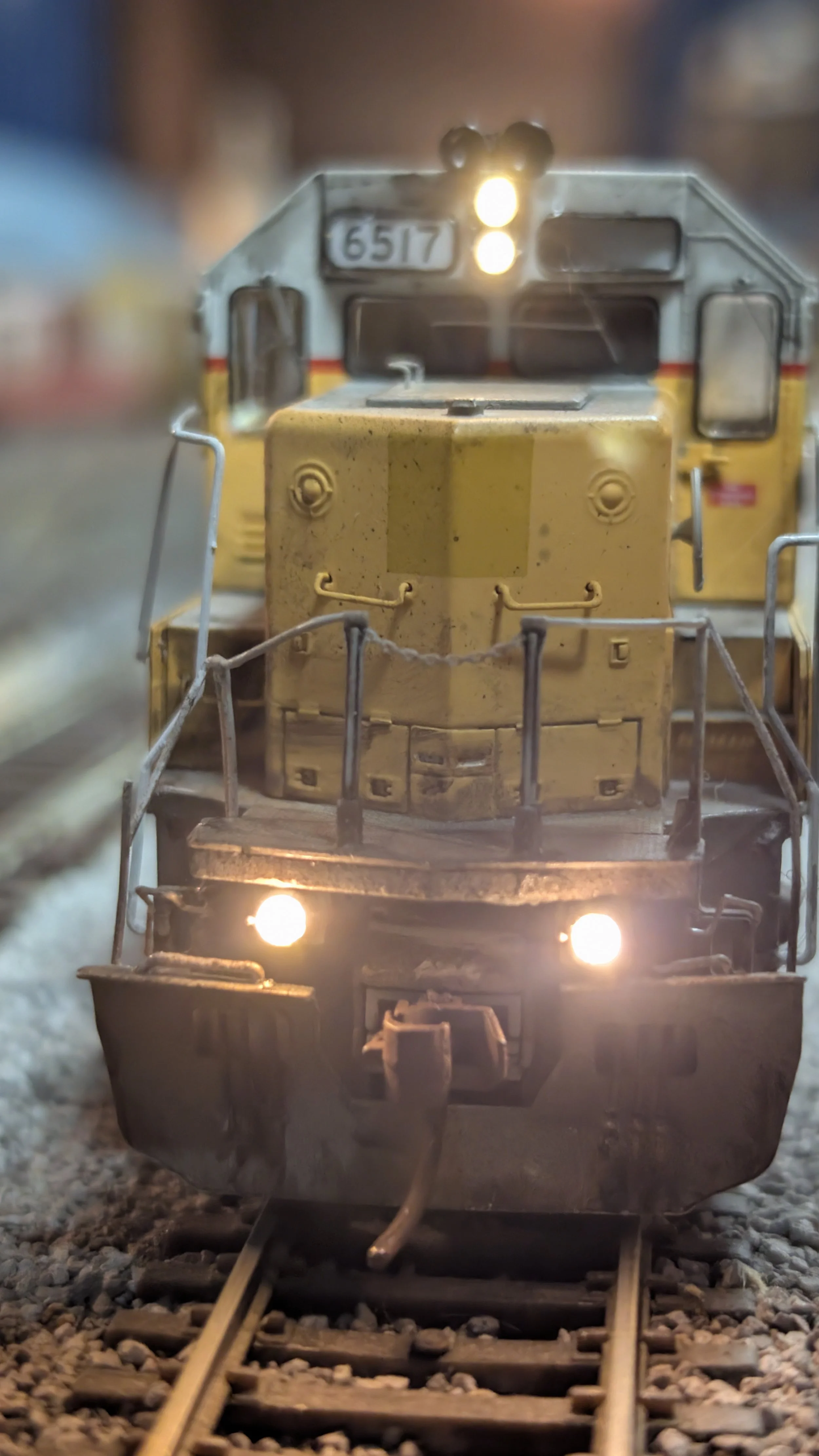 Close-up of a model train engine on a railway track, with front lights on and a blurred background.