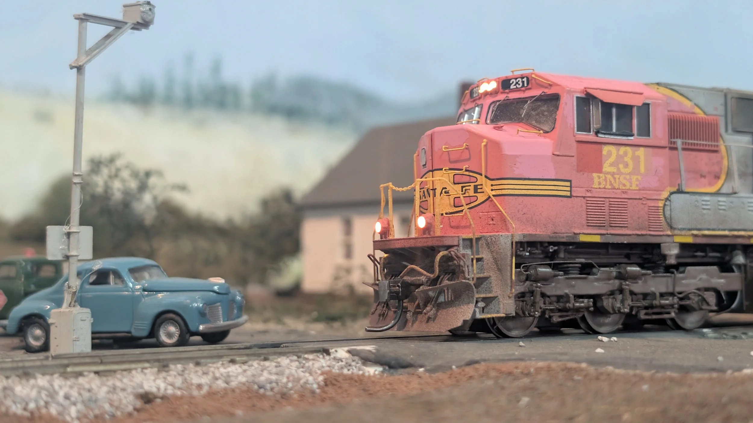 Close-up of a red and yellow train locomotive with the number 231 on the front, passing through a model town with a blue vintage car, a traffic signal, and buildings in the background.