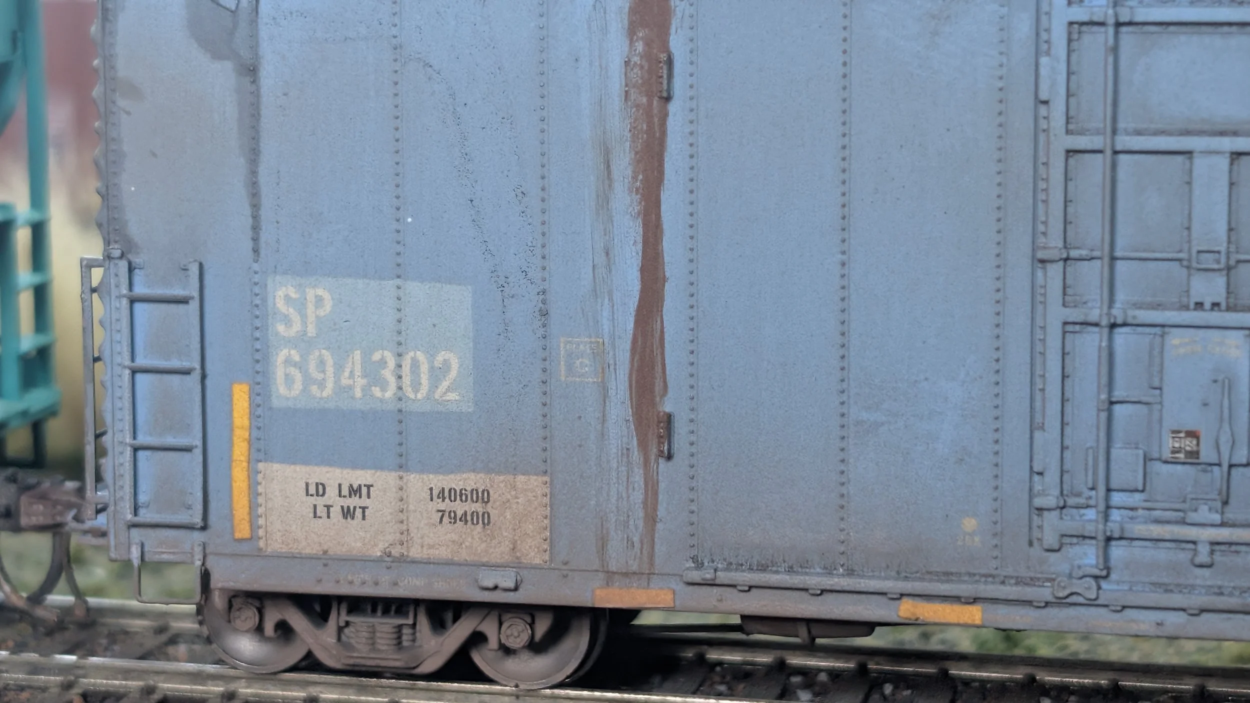 Close-up of a blue model train car with identification numbers and labels, showing weathering details like rust and dirt, on a railway track.