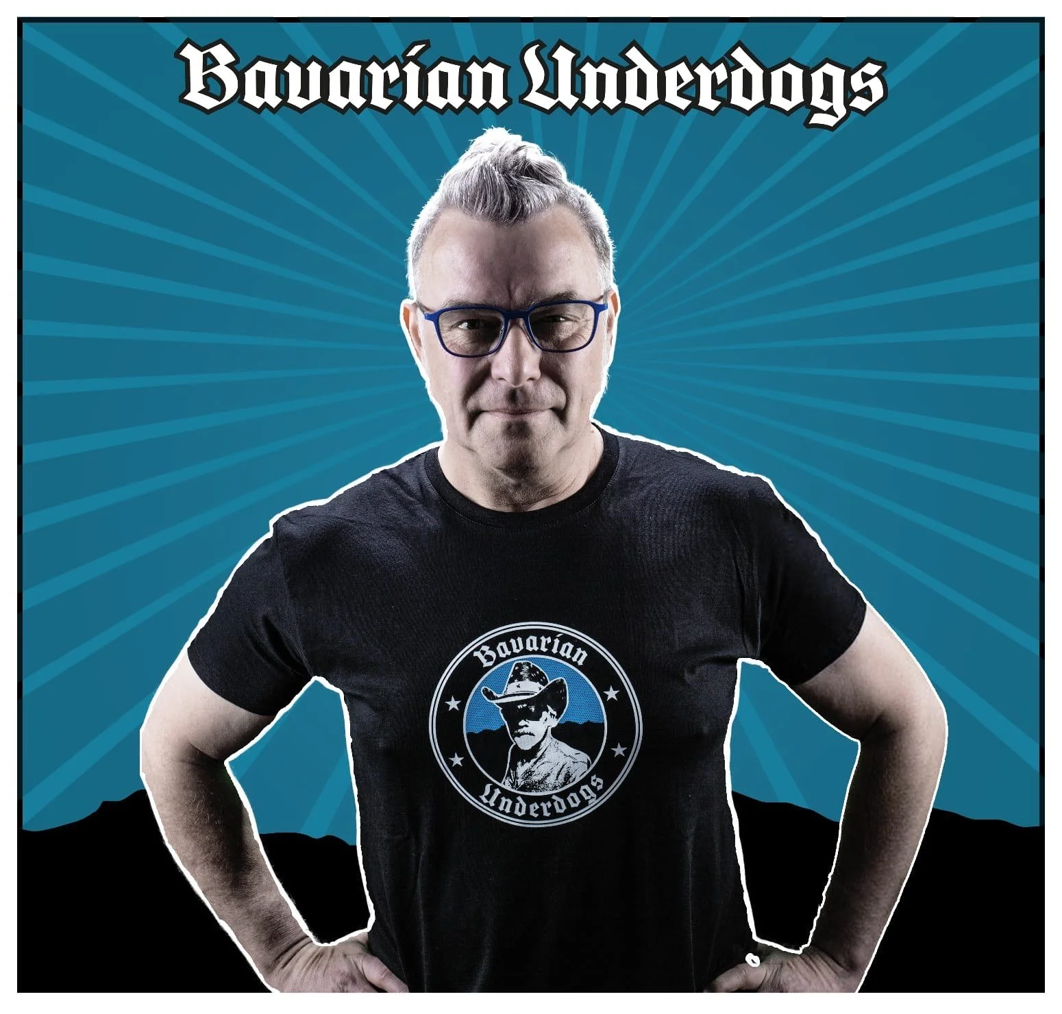 Lucky Zapatta mit Brille und kurzem, blondem Haar, trägt ein schwarzes T-Shirt mit einem Logo, das einen Mann im Cowboy-Hut zeigt, mit Text 'Bavarian Underdogs'. Er steht vor einem blauen Hintergrund mit radialen Linien.