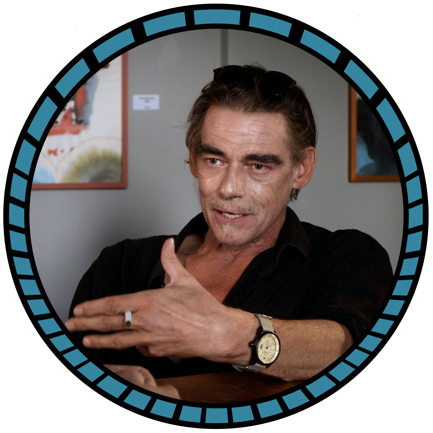 Ein Mann mit dunklen, gewellten Haaren, Brille auf dem Kopf, trägt eine schwarze Hemd und eine Armbanduhr, sitzt an einem Tisch und hält eine Zigarette in der Hand, im Hintergrund sind Bilder an der Wand.