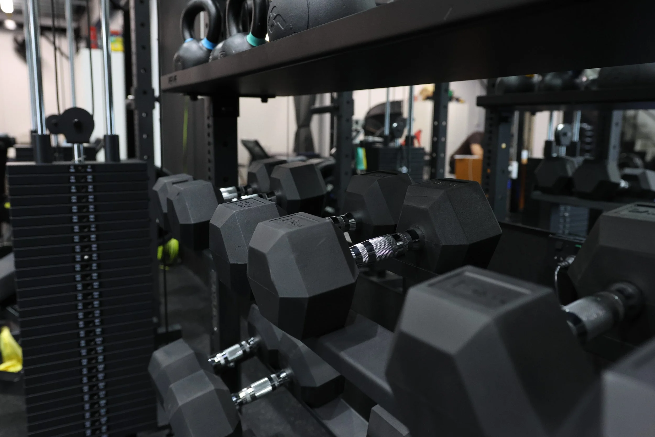 Empty gym with weightlifting equipment, barbells, weight plates, kettlebells, benches, and a blue rubberized workout area.