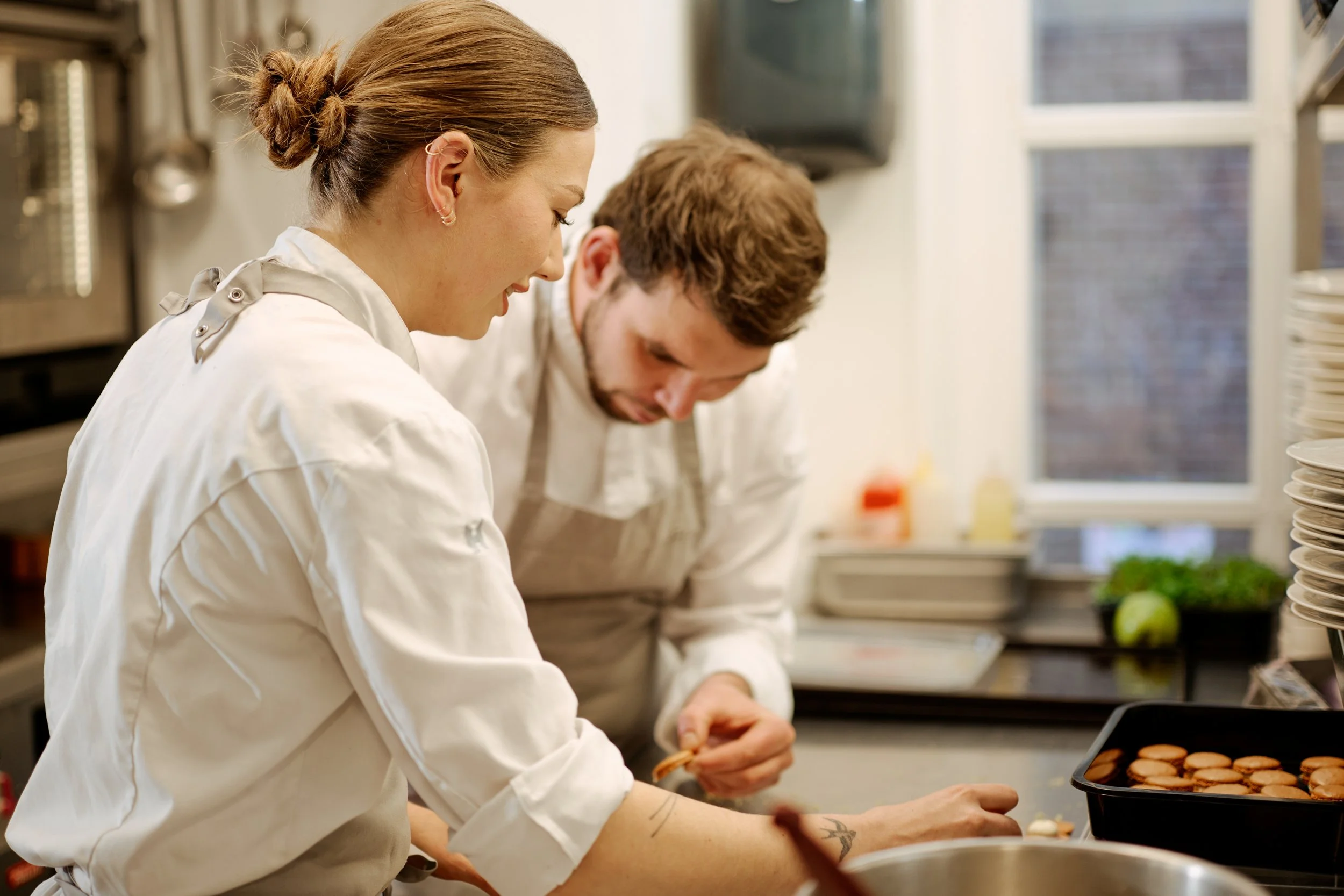 Female and male chefs preparing food in a professional kitchen. The woman is smiling and focusing on her task, while the man is also working intently. There are stacks of plates and baking trays with cookies in the foreground.