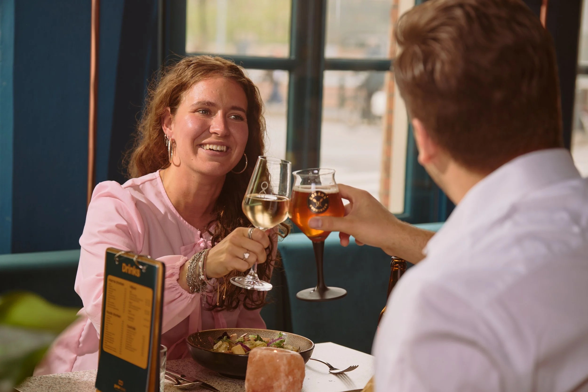 A woman with red hair, wearing a pink top, smiling, holding a glass of beer, toasting with a man who is partially visible, inside a restaurant with large windows and a blue wall.