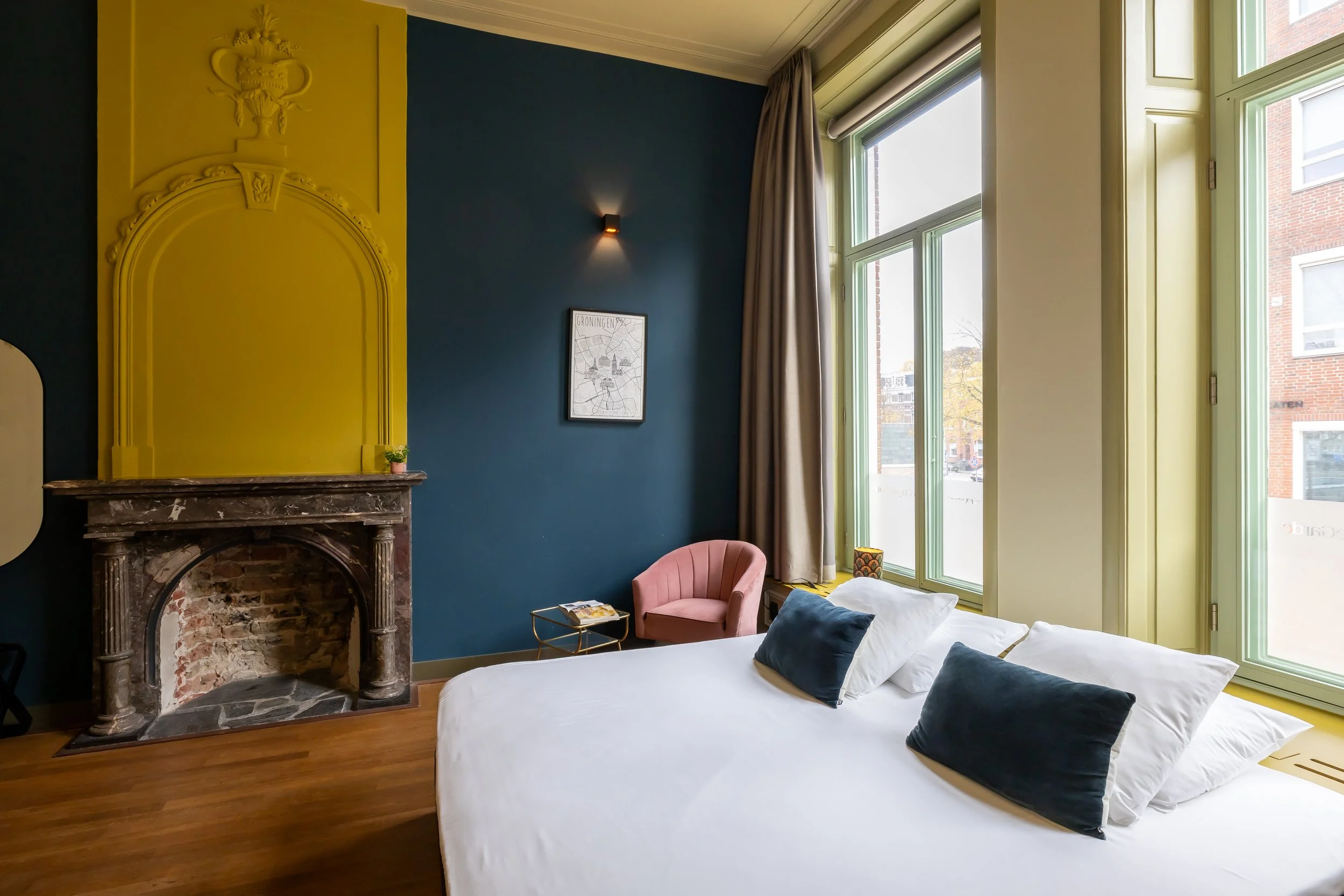 Een helder ingerichte slaapkamer met een groot bed, witte beddengoed met meerdere kussens, een paar fauteuils, grote ramen met beige gordijnen, een muur in donkerblauwe kleur, een schilderij en een vintage open haard met een zwarte marmeren omlijsting.